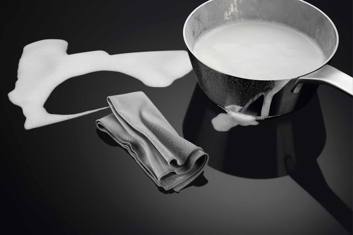 Een pan op een zwart fornuis met overkokende melk. Een grijze doek in de buurt.