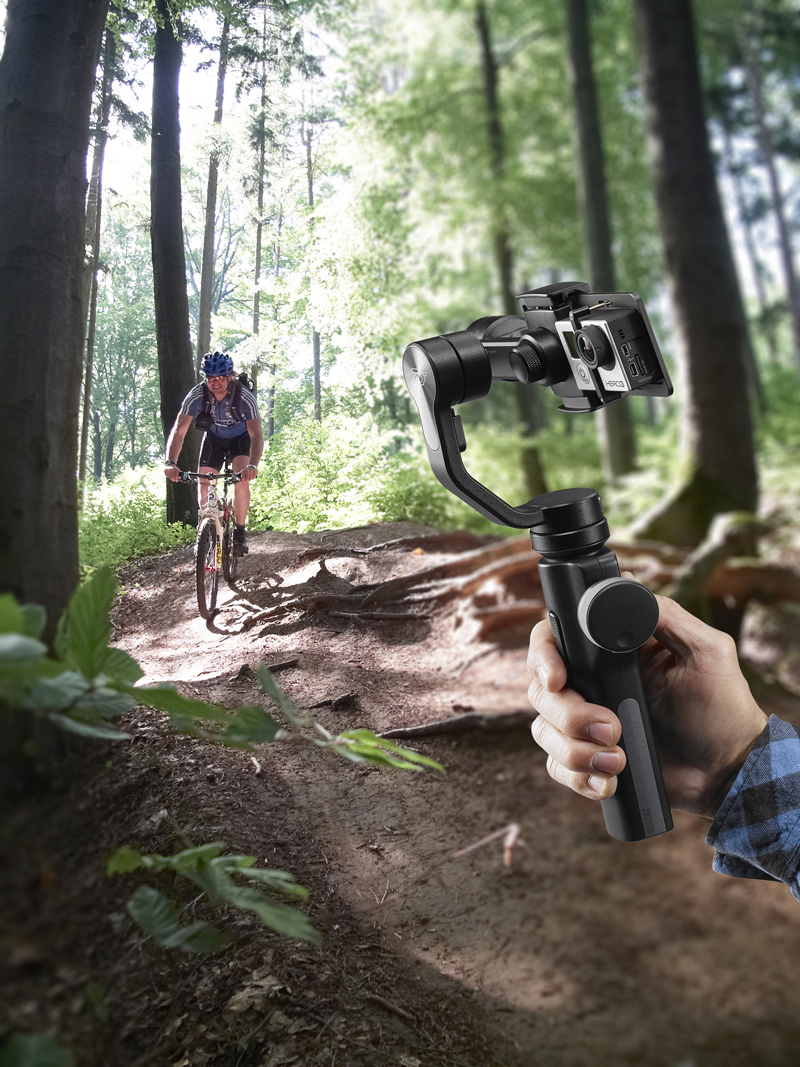 Eine Hand hält einen Kamerastabilisator in einem Wald und filmt einen Mountainbiker auf einem Waldweg.