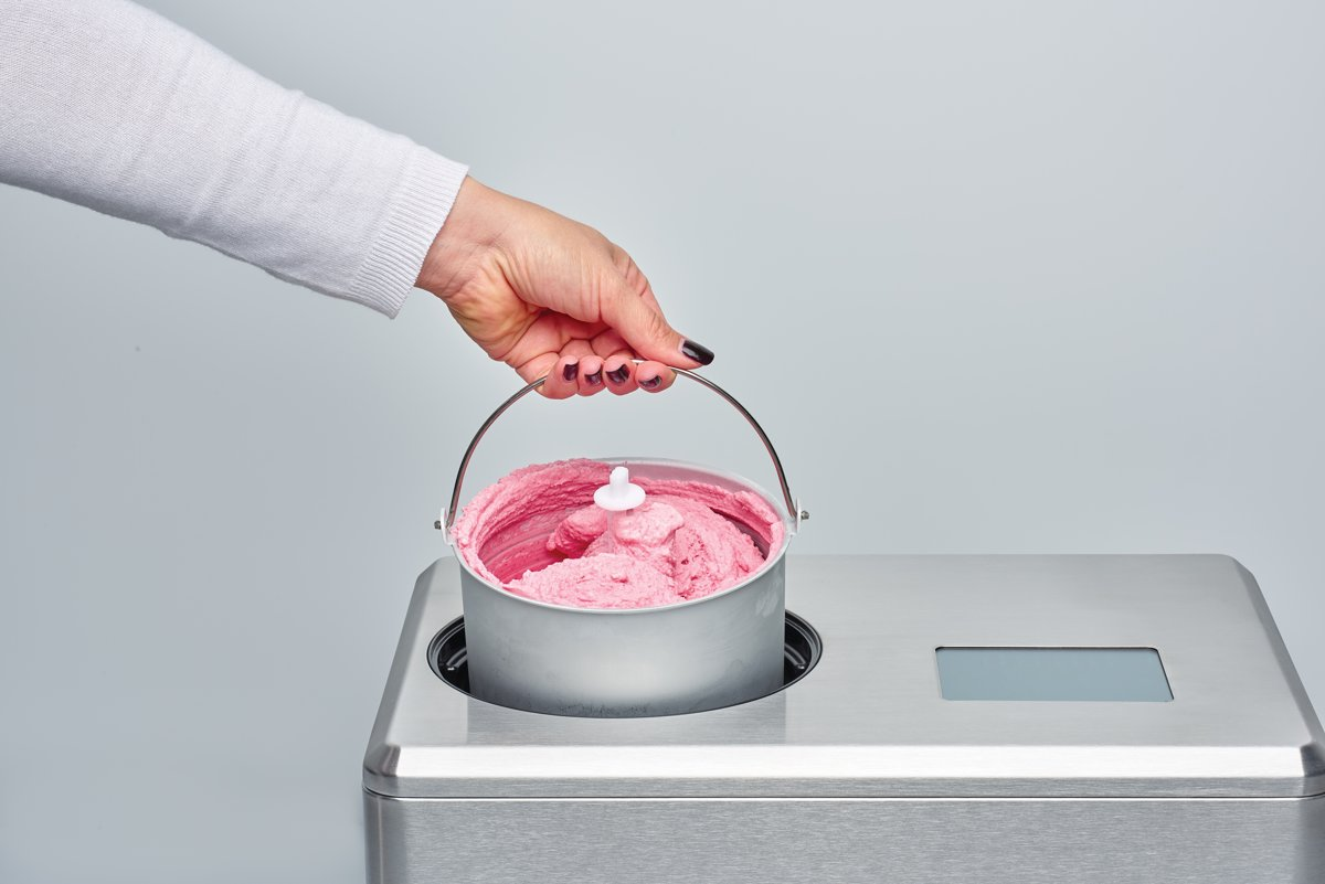 Een hand die een metalen kom met roze ijs vasthoudt boven een ijsmachine, op wit.