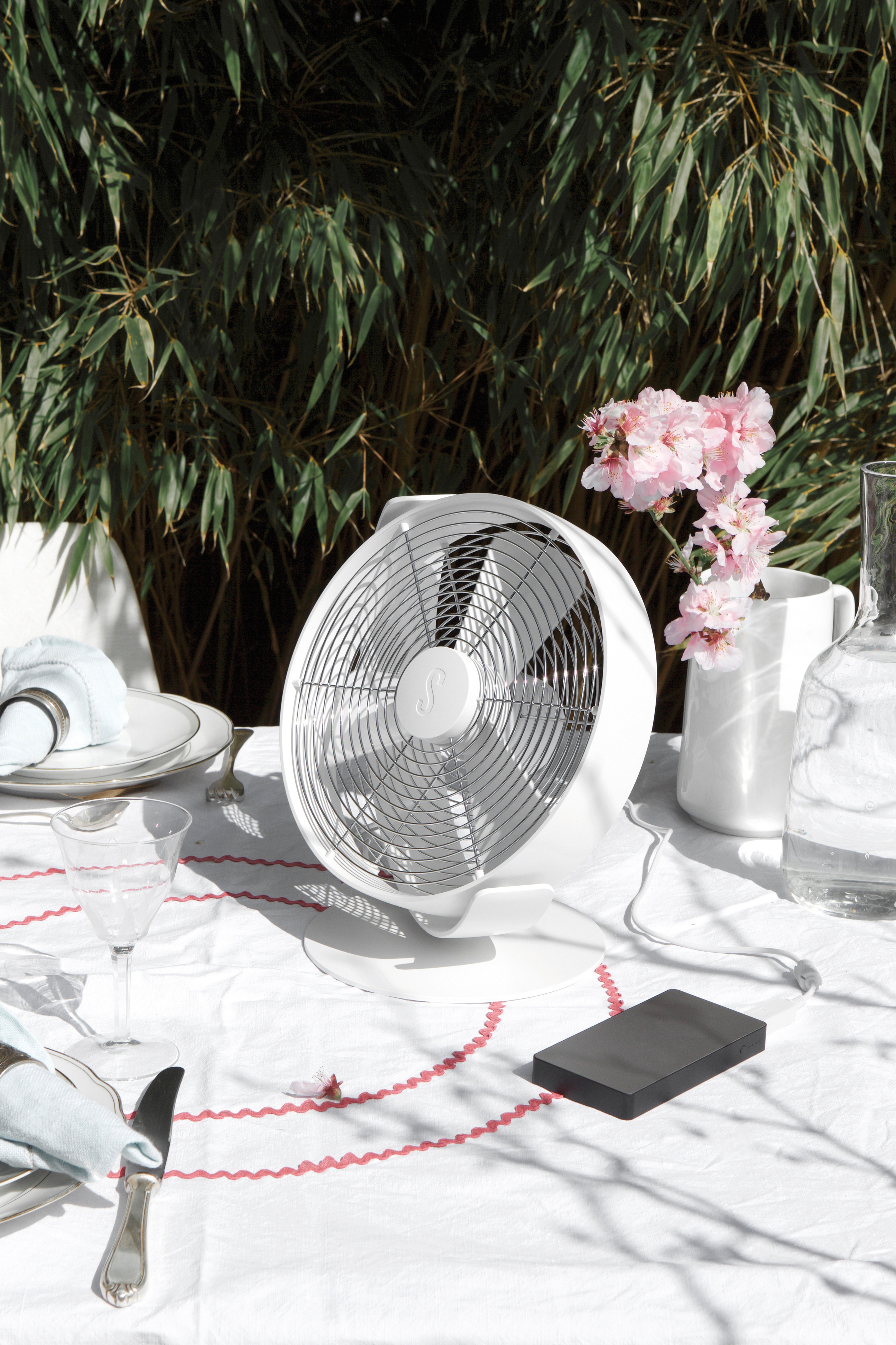 Ein weißer elektrischer Ventilator auf einem weißen Tisch. Auf dem Tisch befinden sich eine schwarze Powerbank, Blumen und Glaswaren.