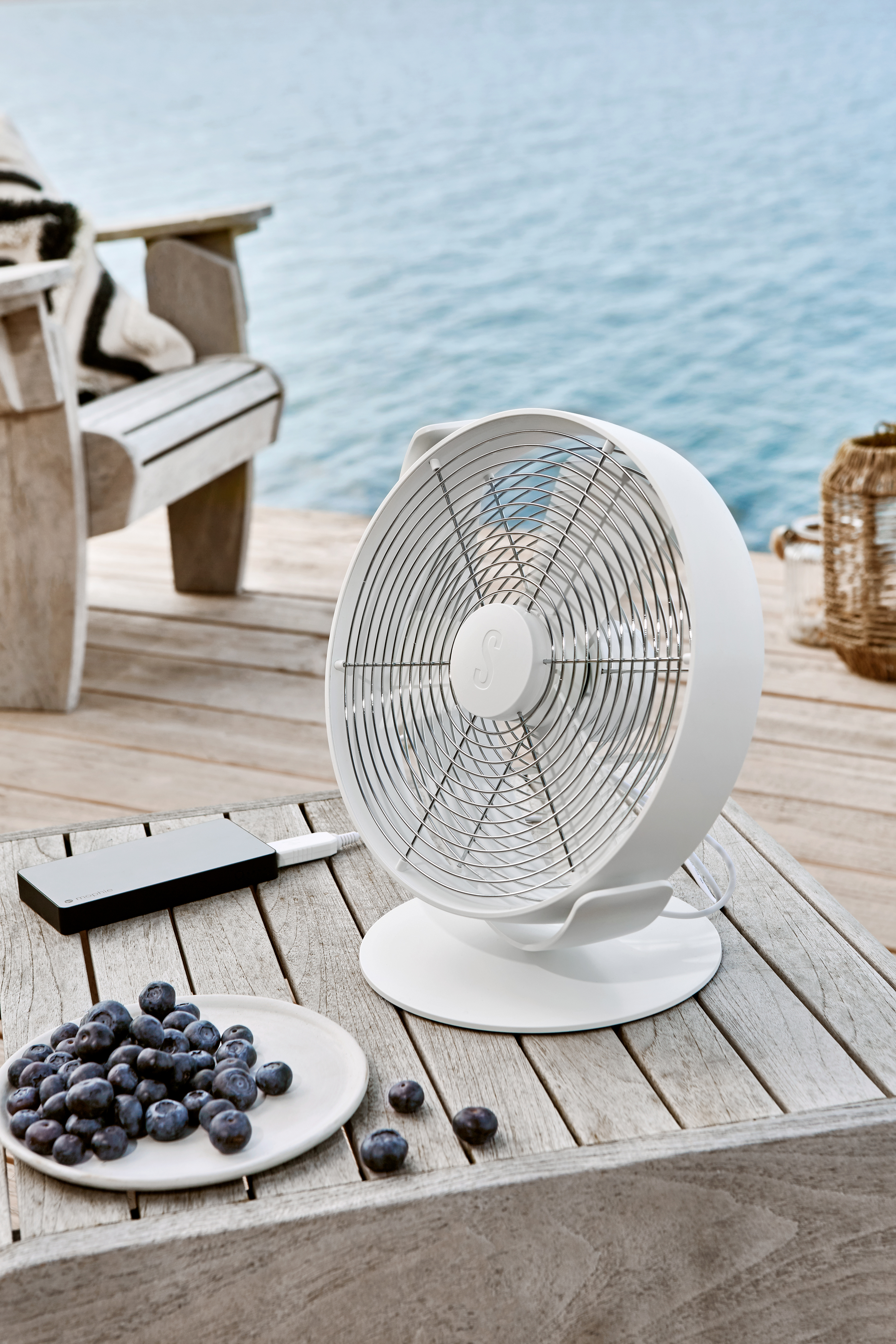 Weißer Ventilator auf Holztisch mit Blaubeeren, Meer im Hintergrund.