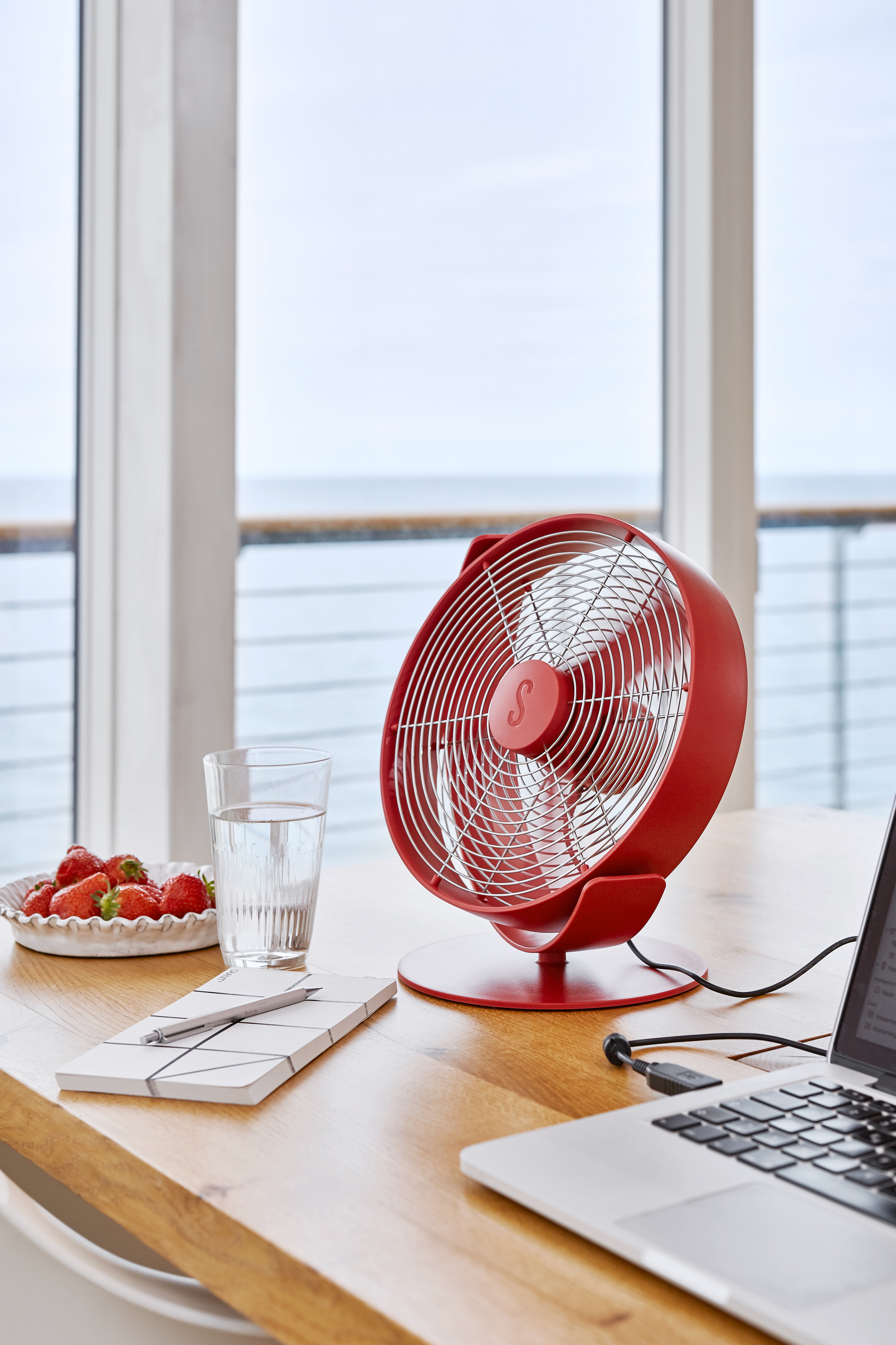 Ein roter Ventilator, Erdbeeren, ein Wasserglas, ein Notizblock und ein Laptop auf einem Holztisch am Fenster.