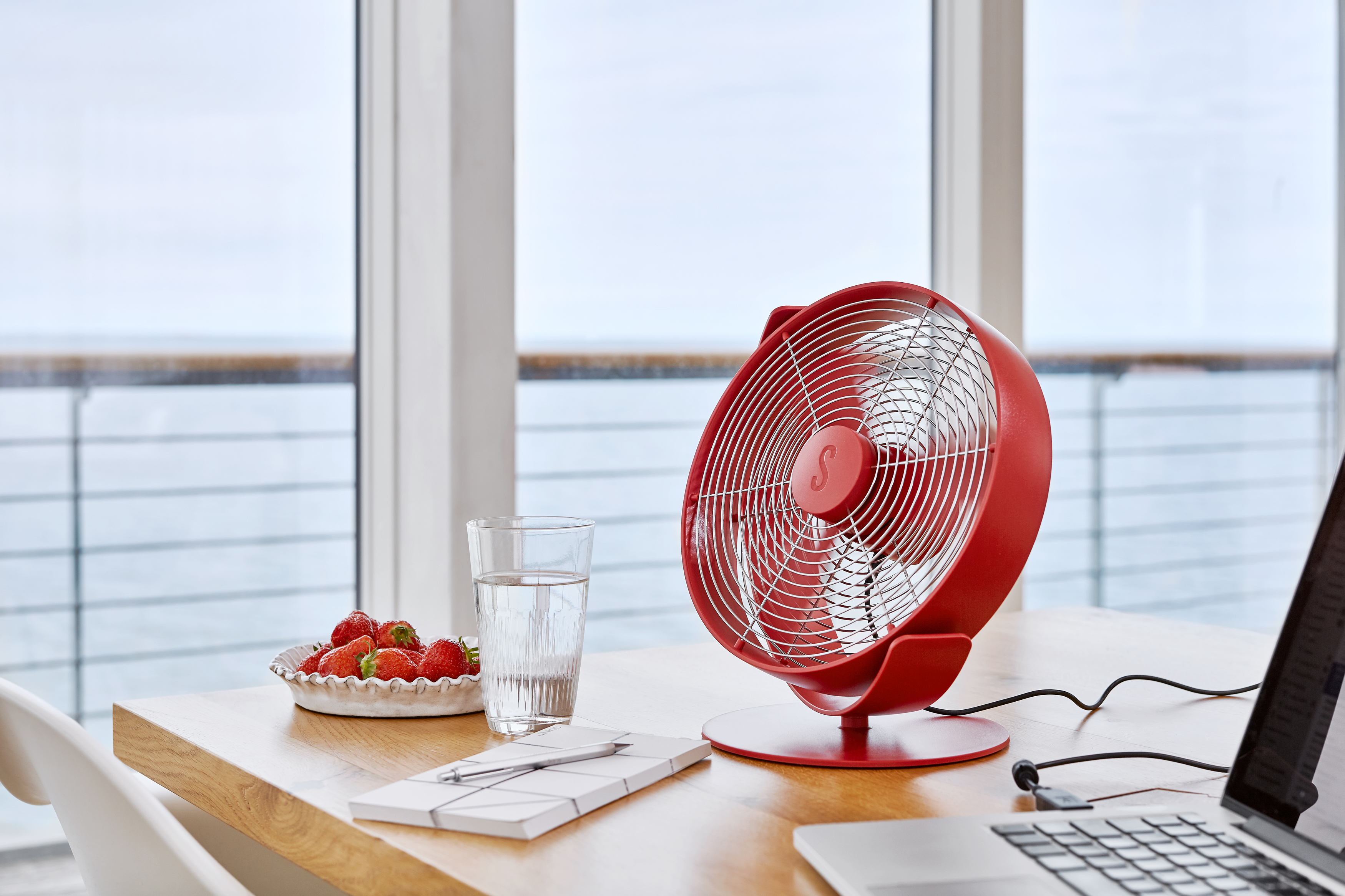 Roter Ventilator auf Tisch, Erdbeeren auf einem Teller, Wasserglas, Notizbuch und Laptop. Fenster und Meer im Hintergrund.