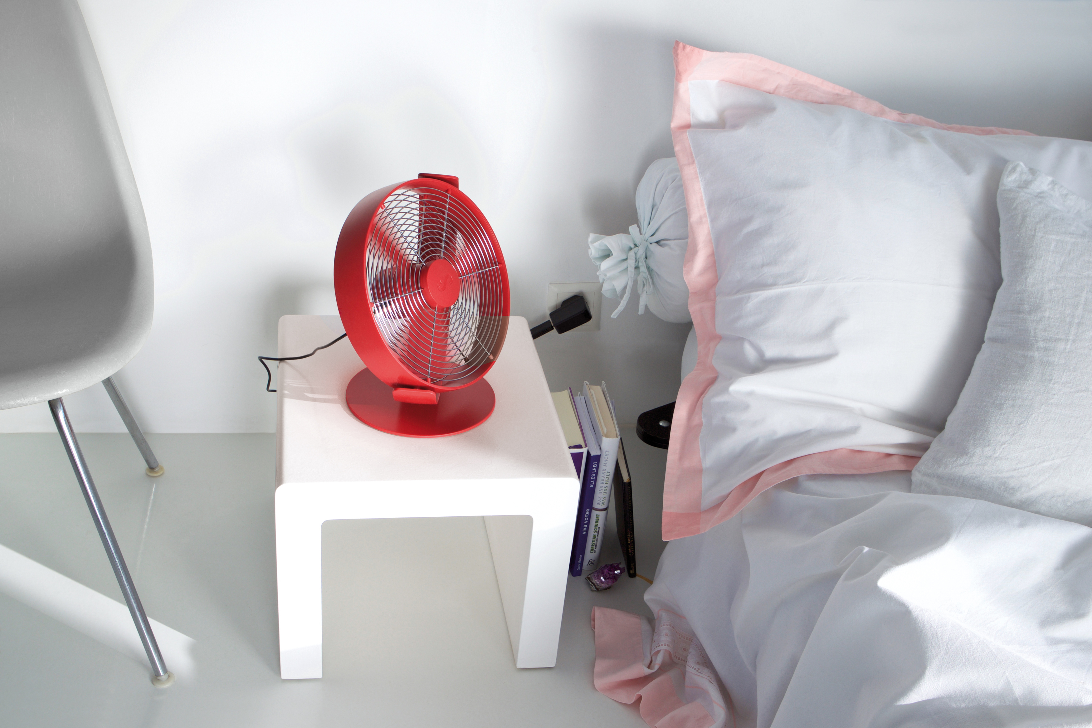 Ein roter Ventilator steht auf einem weißen Tisch neben einem Bett mit weißer Bettwäsche und einem rosa Rand. Im Hintergrund ein grauer Stuhl.