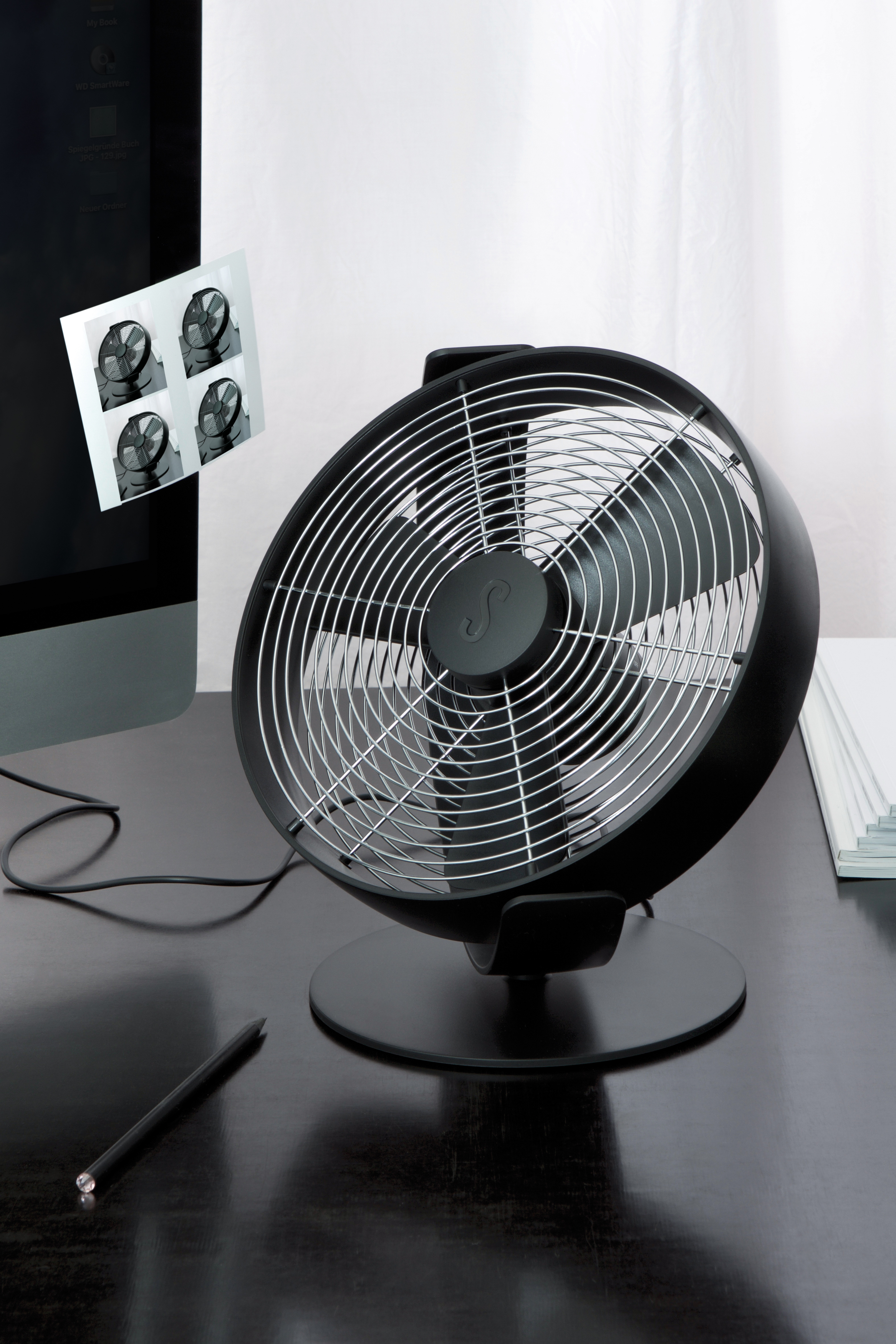 Schwarzer Schreibtischventilator mit schwarzem Sockel. Ein Monitor zeigt Lüfterbilder. Auf dem Schreibtisch liegt ein Stift.
