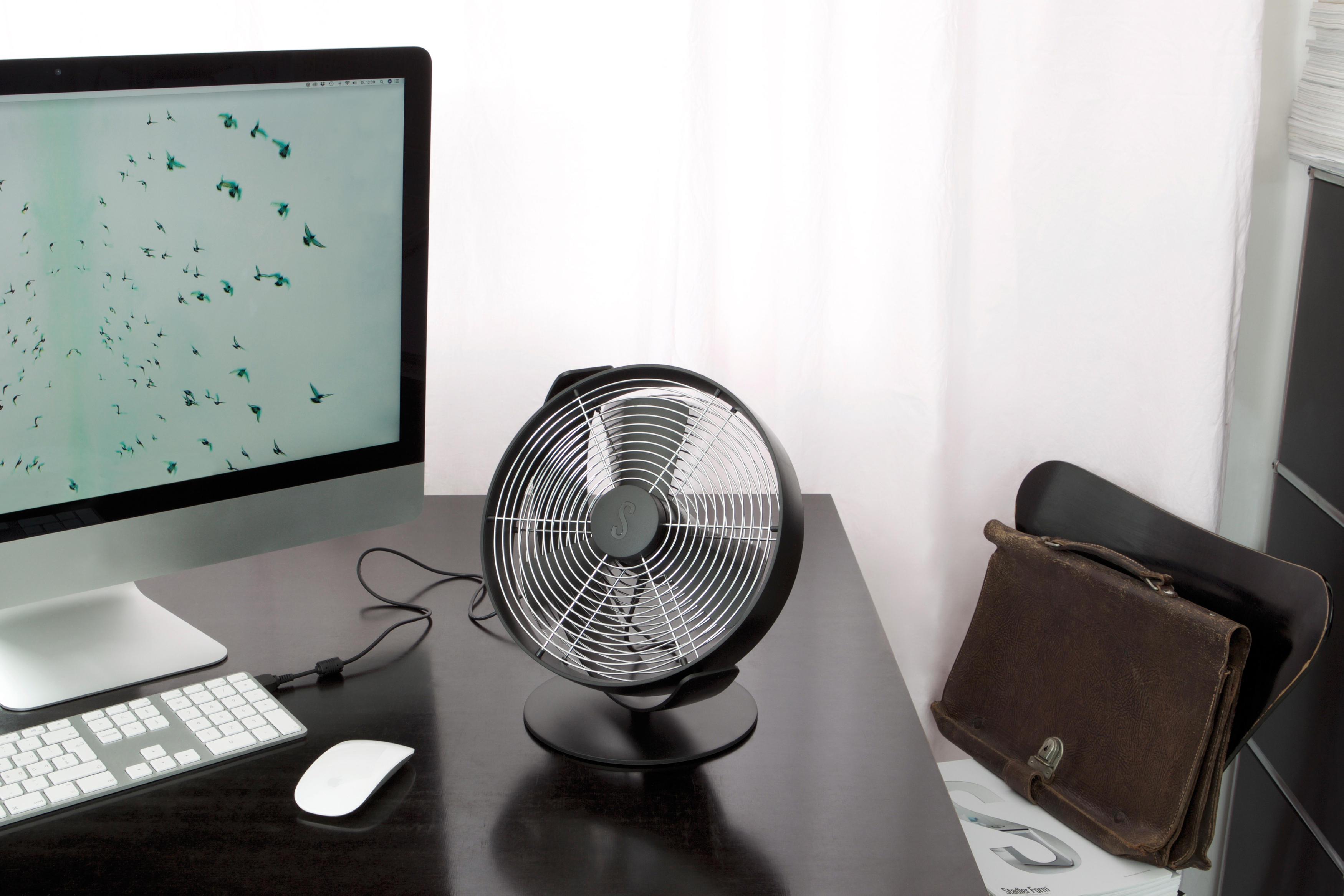 Schwarzer Ventilator auf dem Schreibtisch. Ein Computermonitor, Tastatur und Aktentasche.