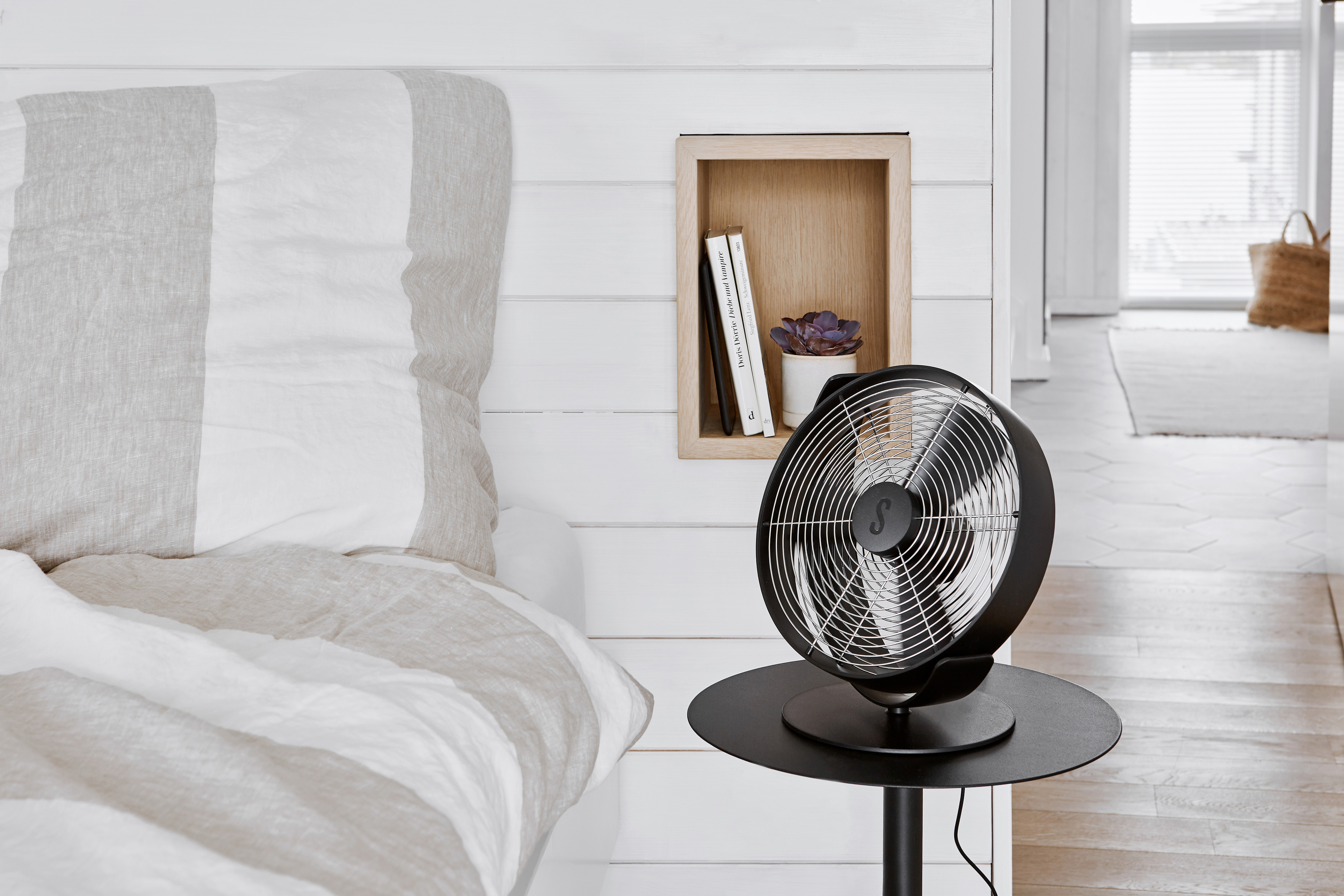 Ein schwarzer Ventilator auf einem Tisch neben einem Bett mit gestreifter Decke. Eine Wand hat eine Nische.