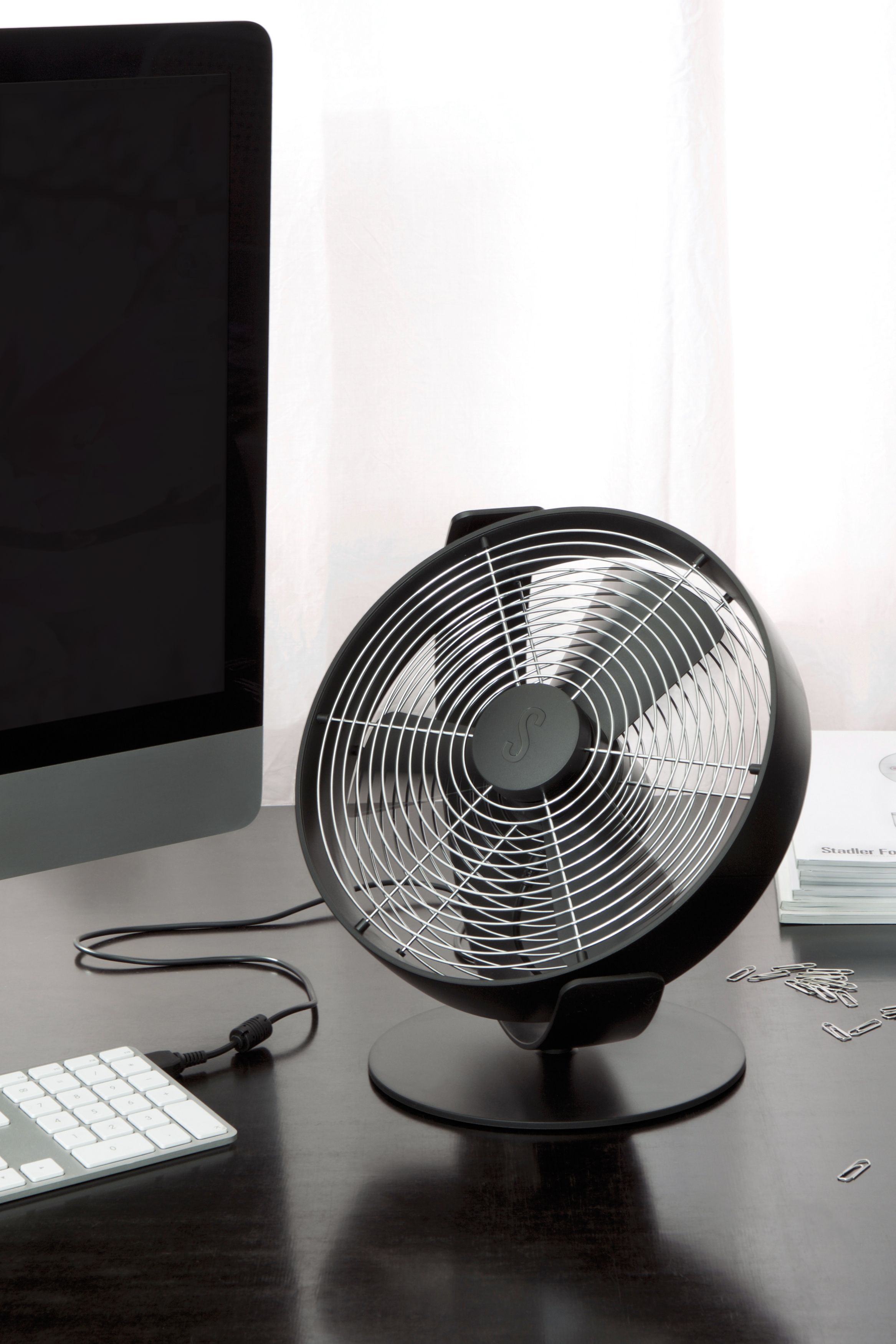 Schwarzer Tischventilator mit silbernem Gitter, neben Monitor und Tastatur.