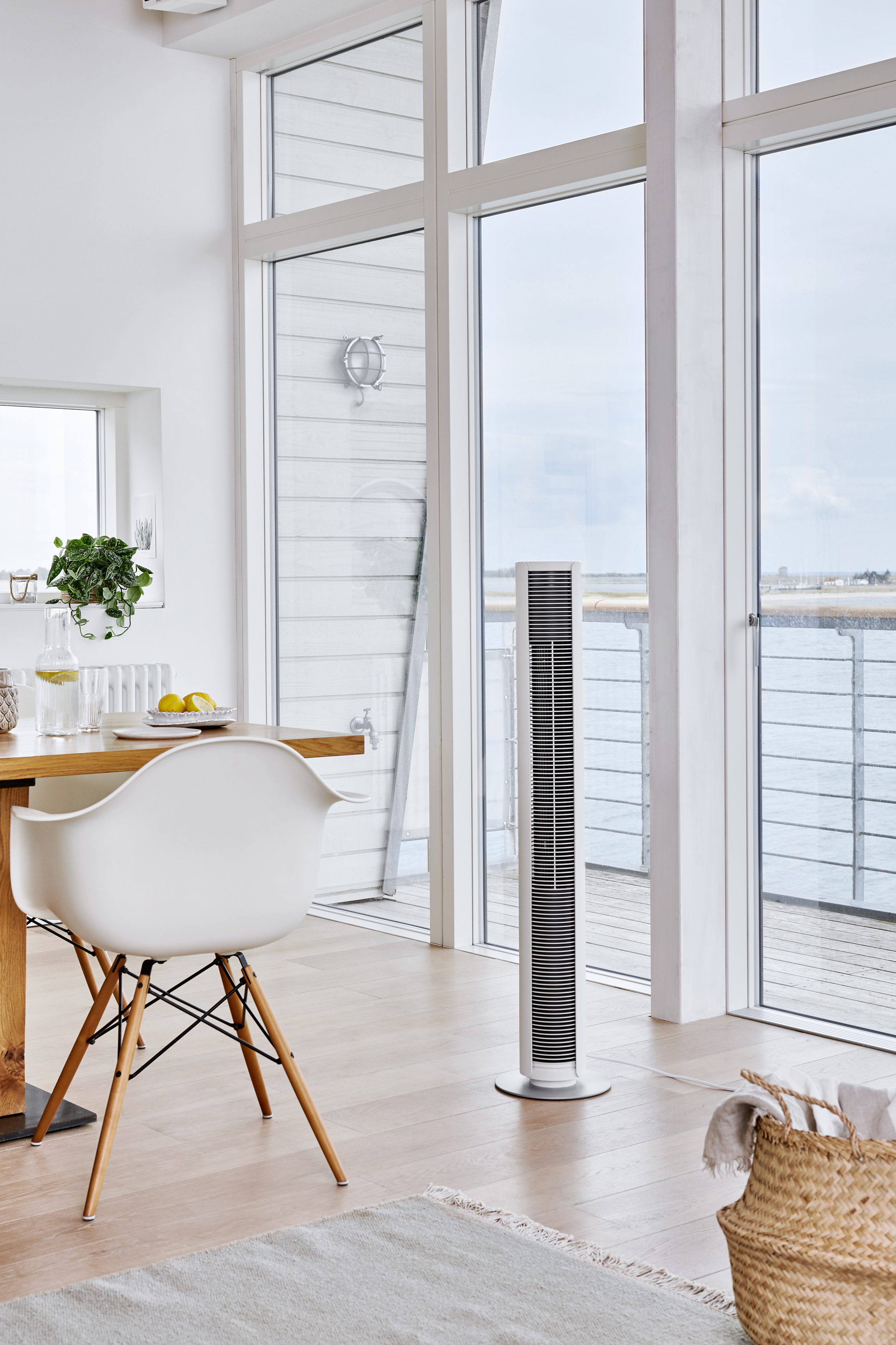 Ein weißer Turmventilator in einem hellen, modernen Raum. Es gibt einen Tisch und einen Stuhl, mit Meerblick.