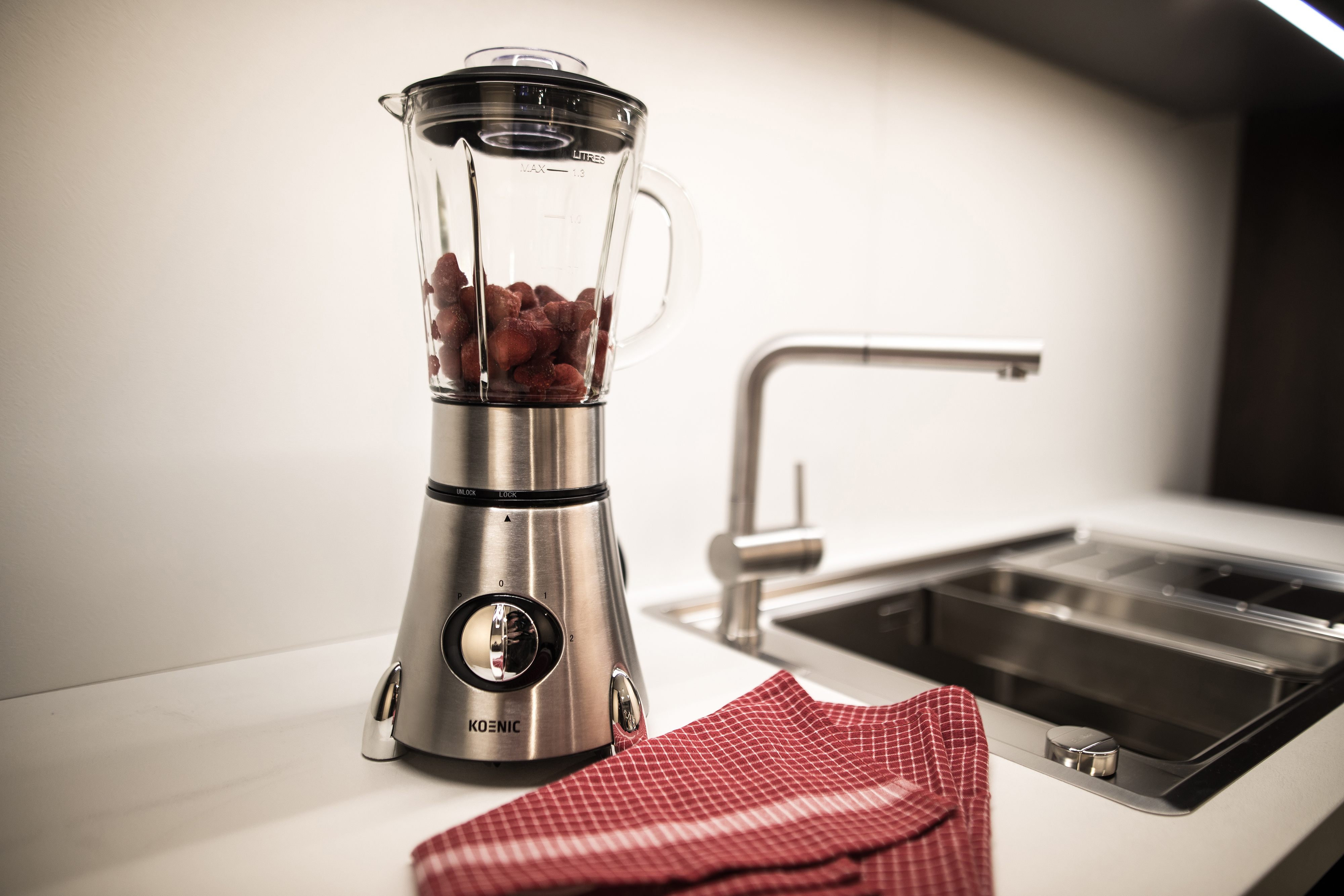 Silberner Mixer mit Erdbeeren, auf einer weißen Arbeitsplatte. Ein Waschbecken und ein rot kariertes Tuch sind vorhanden.