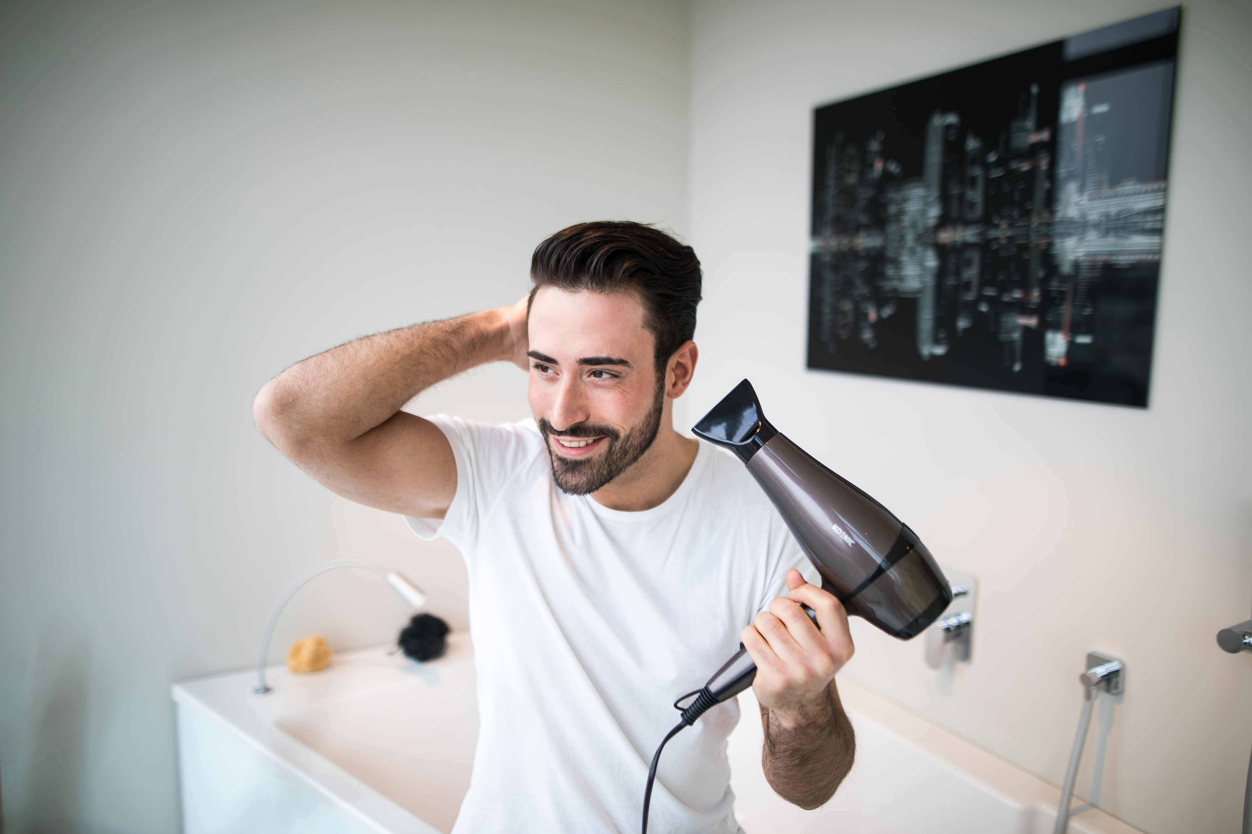 Ein Mann im Badezimmer trocknet seine Haare mit einem Fön. Er lächelt, blickt in einen Spiegel, weißer Hintergrund.