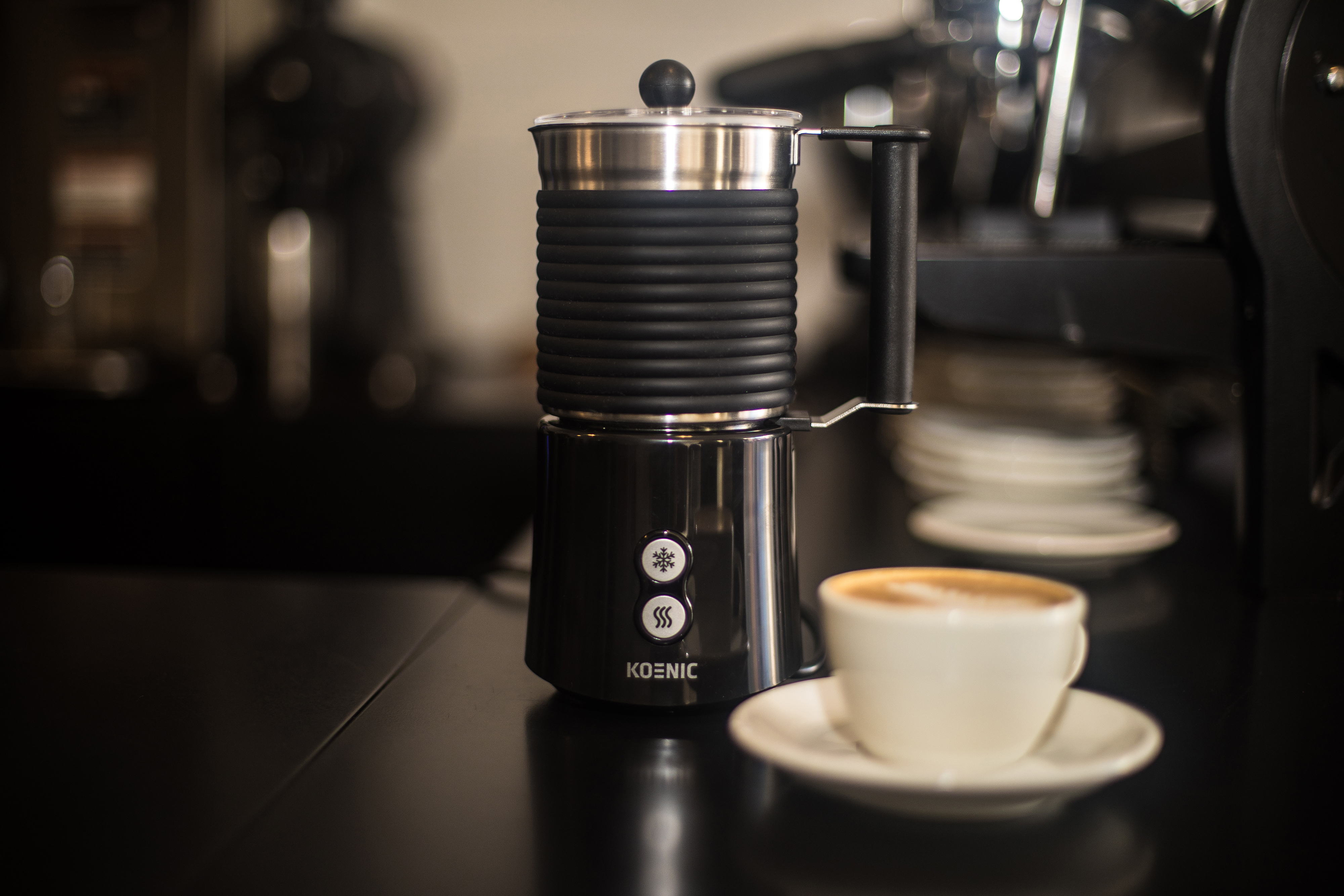 Ein schwarzer Milchaufschäumer und eine Tasse Kaffee auf einer schwarzen Oberfläche. Verschwommener Hintergrund mit Regalen.
