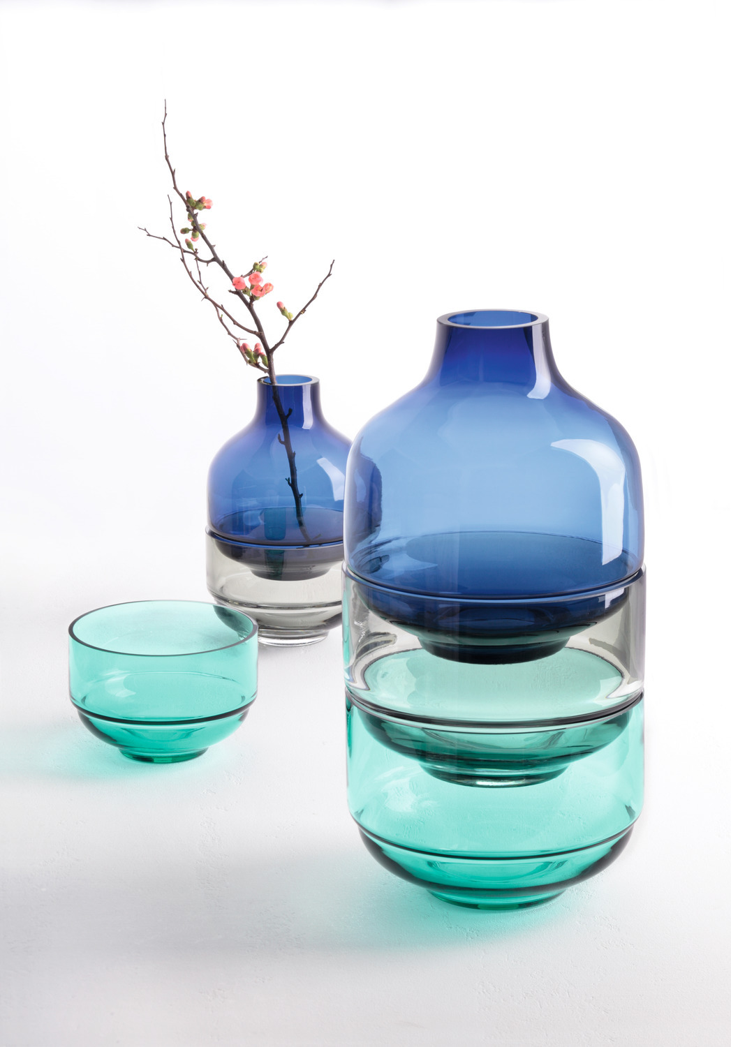 Drei blaue und tealfarbene Glasvasen, eine mit einem Blumenast, auf weißem Hintergrund.