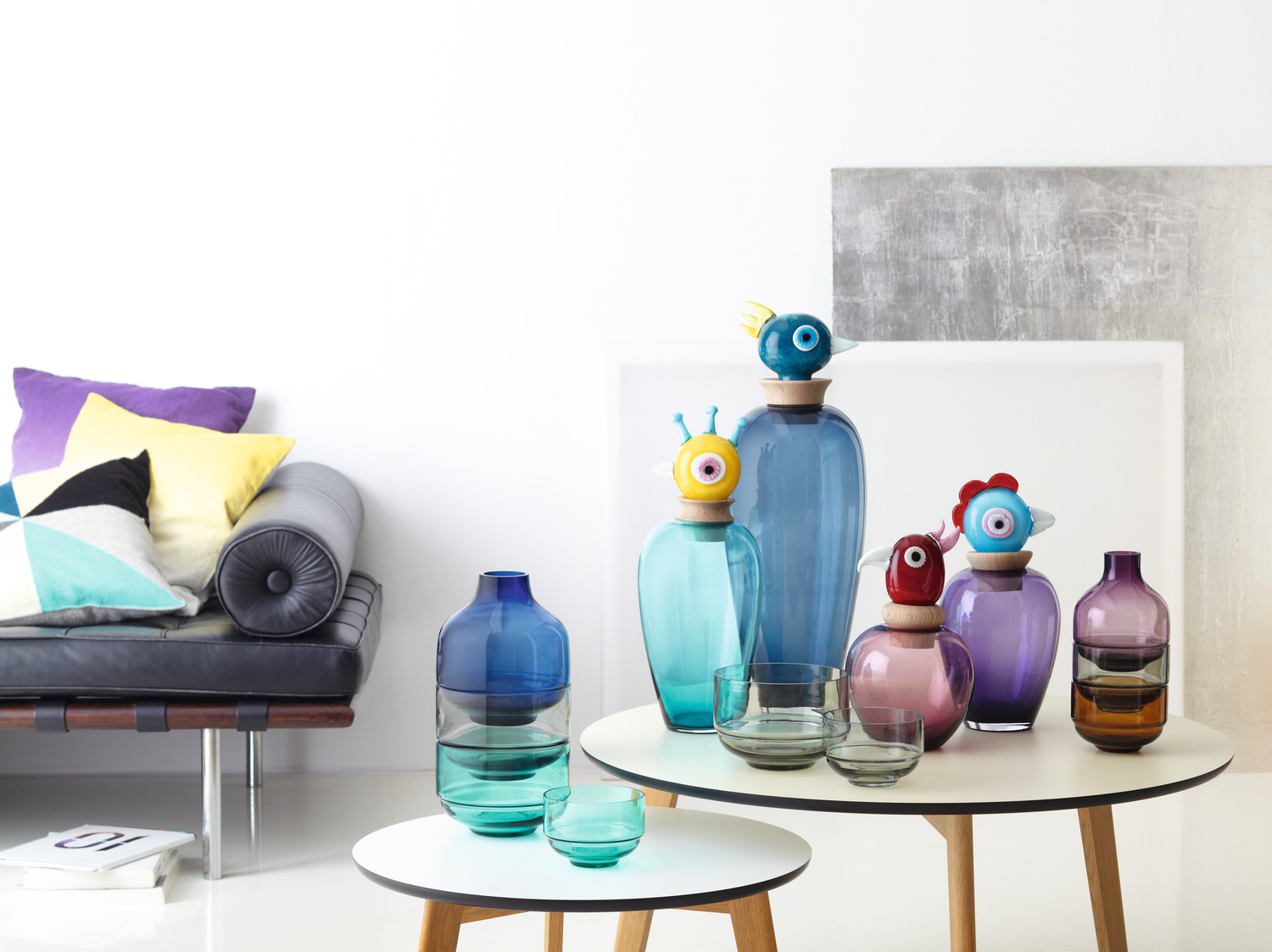 Sammlung bunter Glasvasen mit dekorativen Aufsätzen auf einem weißen Tisch.