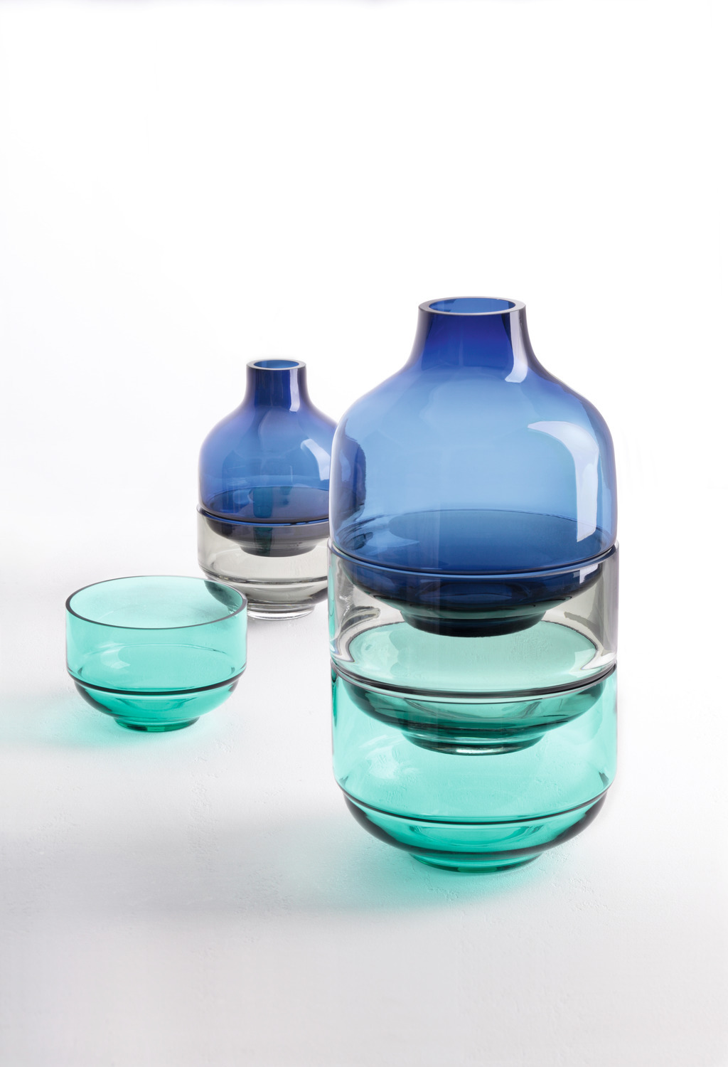 Drei gestapelte Glasvasen in Blau und Türkis. Ein kleines türkisfarbenes Glas daneben.