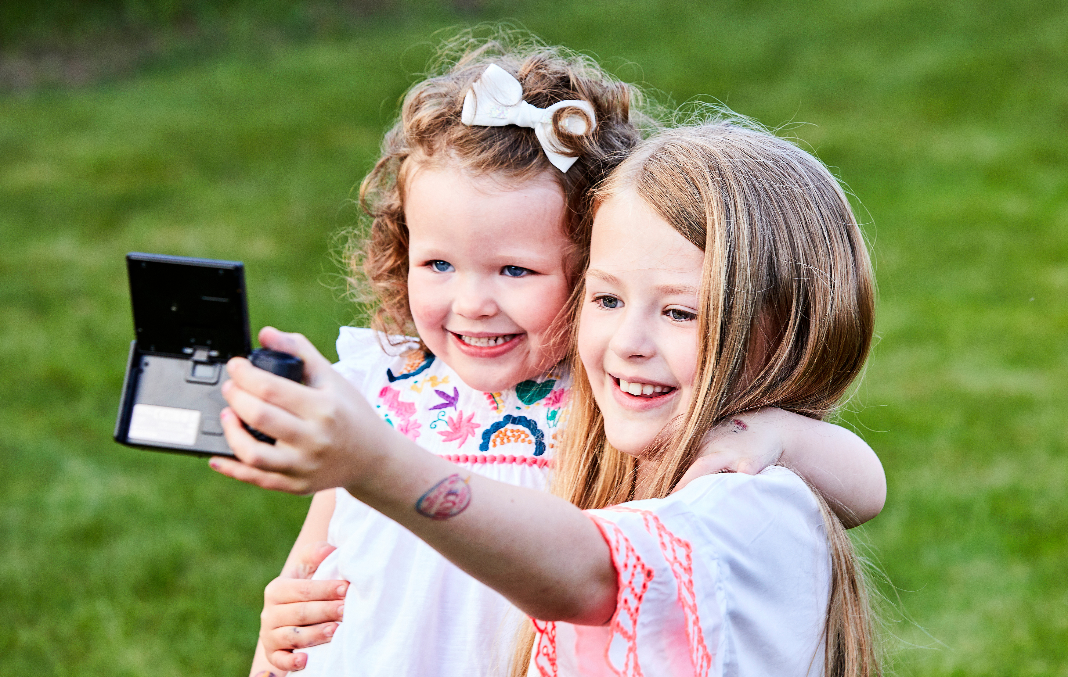 Zwei junge Mädchen machen ein Selfie mit einer Kamera und lächeln in einer grünen, grasbewachsenen Umgebung.