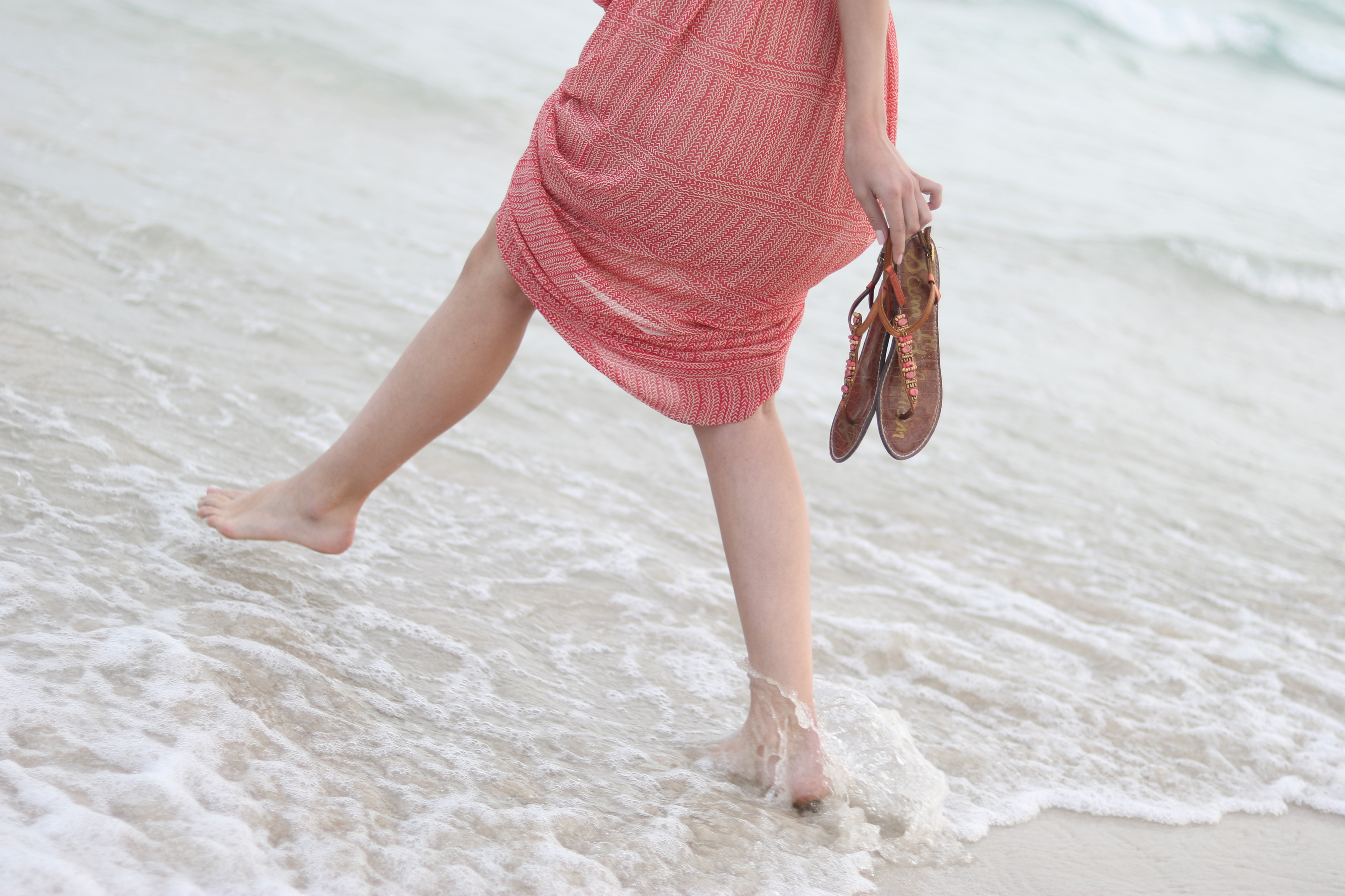 Eine Frau, die am Strand spaziert und Sandalen hält. Rotes Kleid.