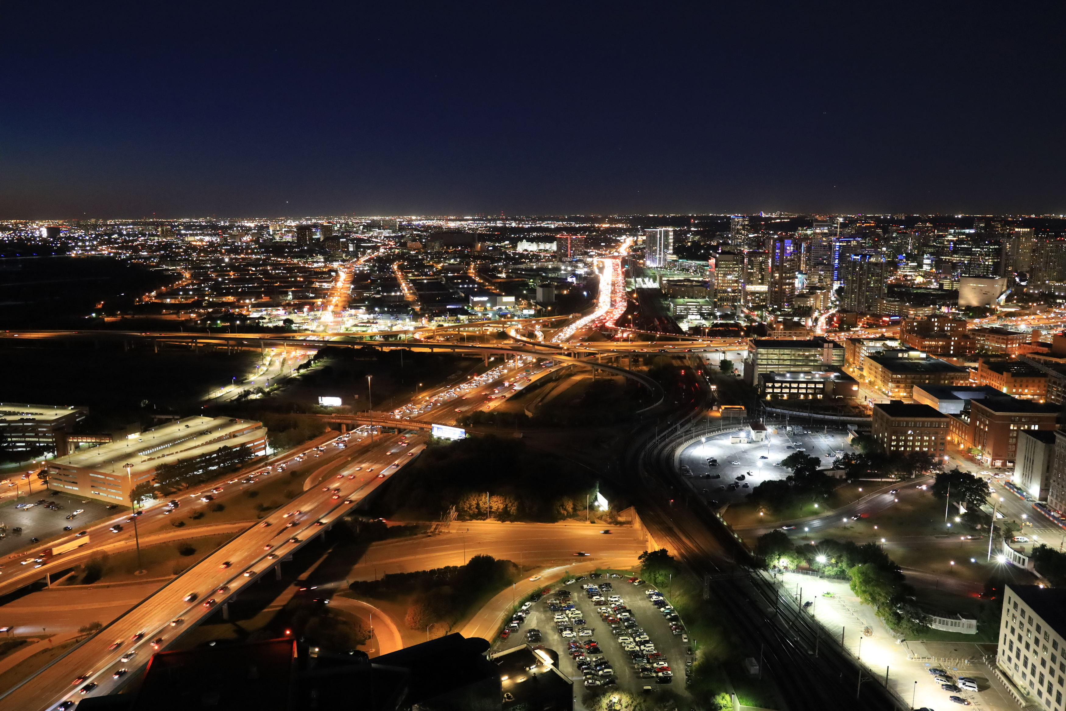 Stadtlandschaft bei Nacht. Lichter von Gebäuden und Straßen. Dunkelblauer Himmel. Ein Parkplatz und eine Überführung sind sichtbar.