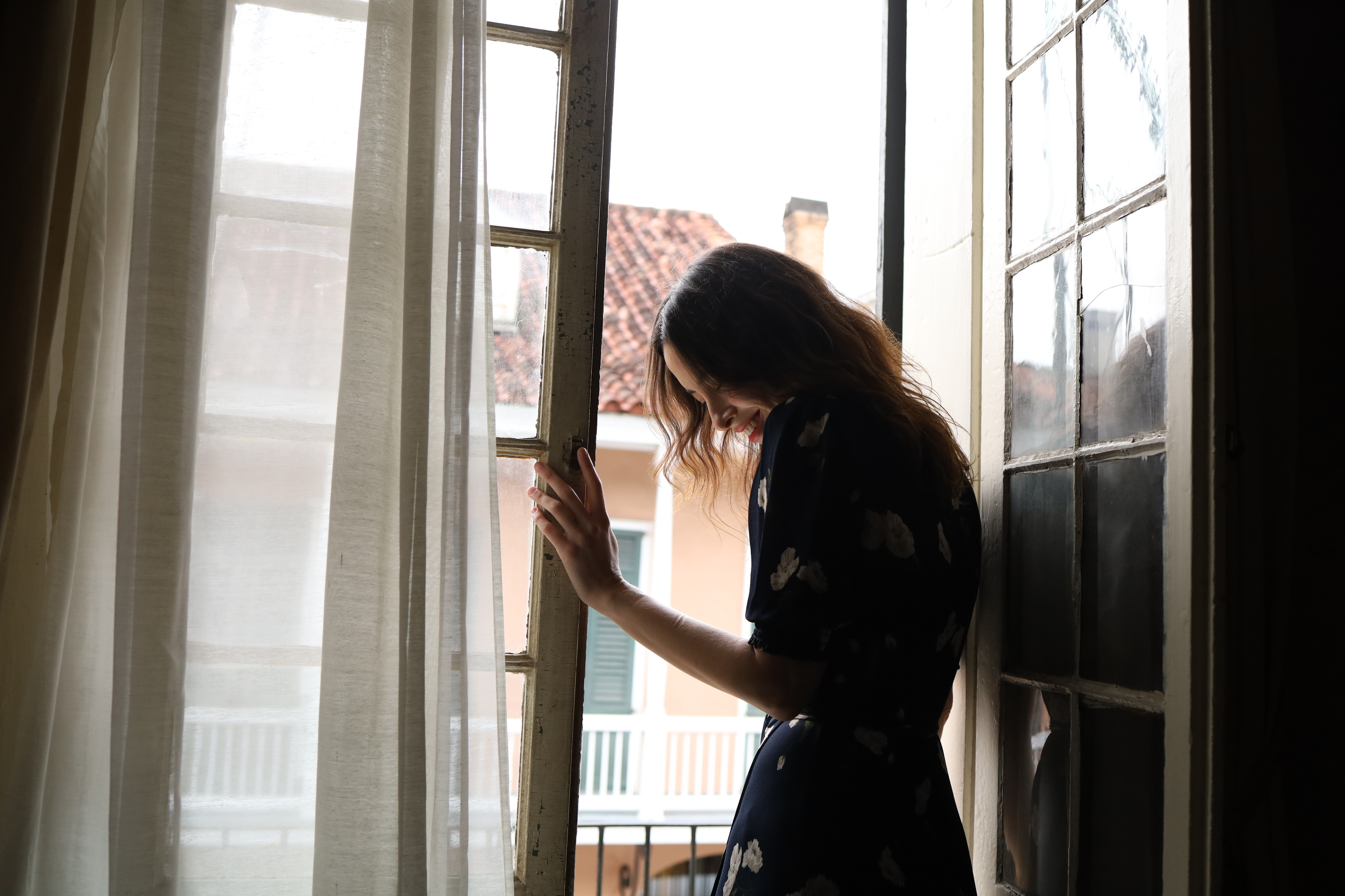 Eine Frau steht an einem offenen Fenster, trägt ein dunkles Kleid und schaut nach unten. Gebäude draußen.