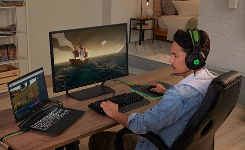 Ein Mann spielt mit Headset und Monitor. Ein Laptop und eine Tastatur befinden sich ebenfalls auf dem Schreibtisch.