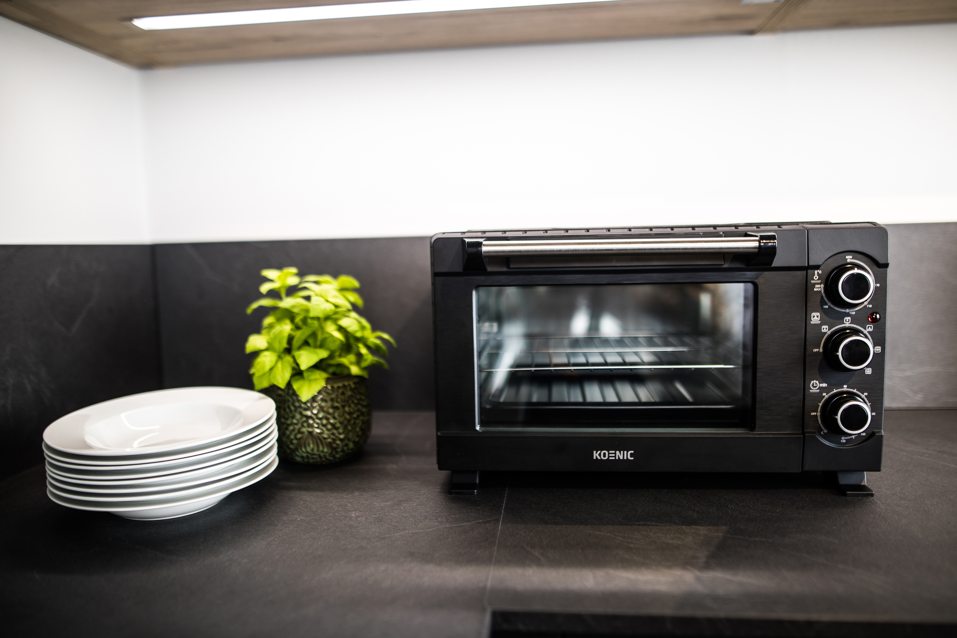 Schwarzer Koenic-Ofen auf Arbeitsplatte, mit Tellern und Pflanze, in Küche.