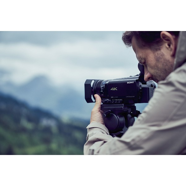 Hombre filmando con una videocámara 4K al aire libre, con montañas al fondo.