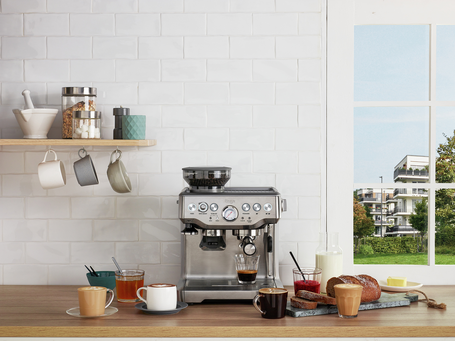 Een roestvrijstalen espressomachine met kopjes, eten en een raam met een groen landschap.