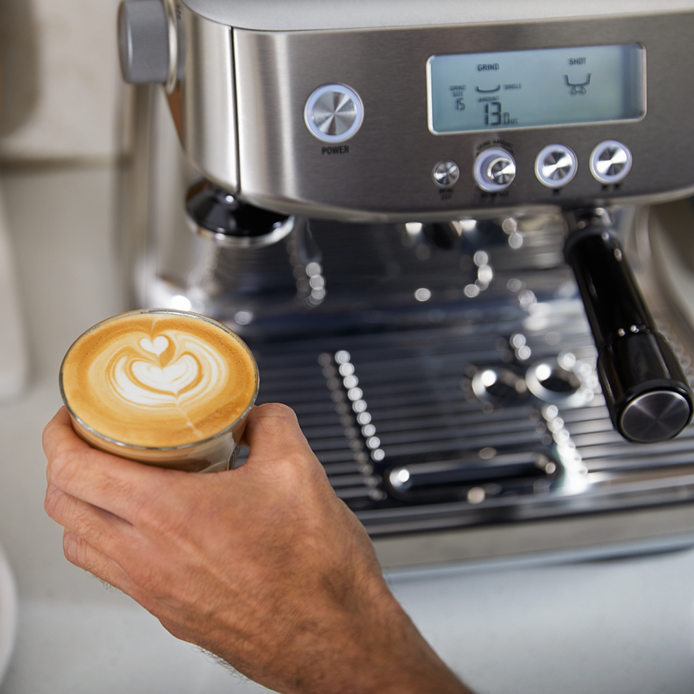 A person holding a glass of latte art next to a silver espresso machine.