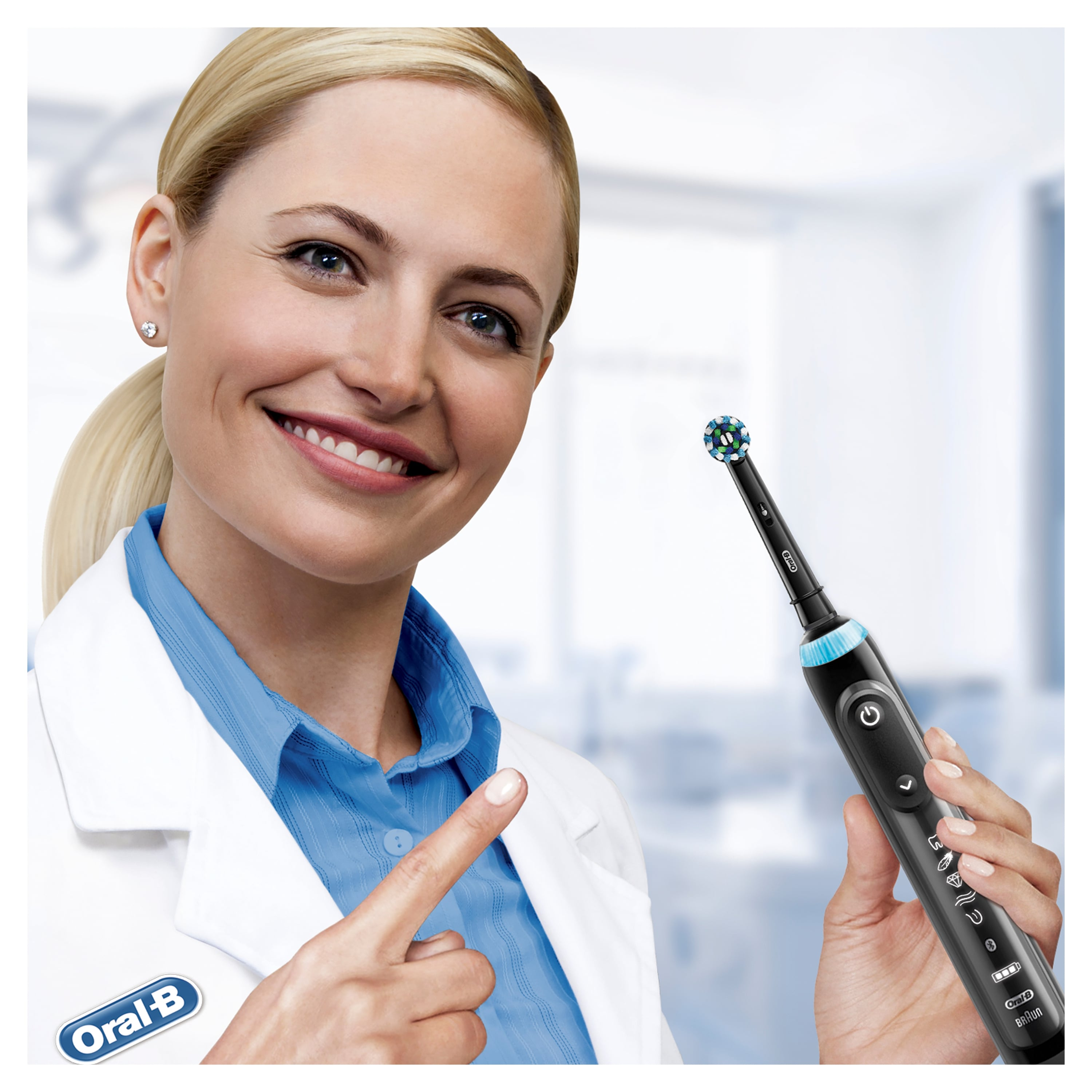 Blonde Frau im Laborkittel zeigt auf eine elektrische Zahnbürste von Oral-B. Lächelt und hält die Bürste.