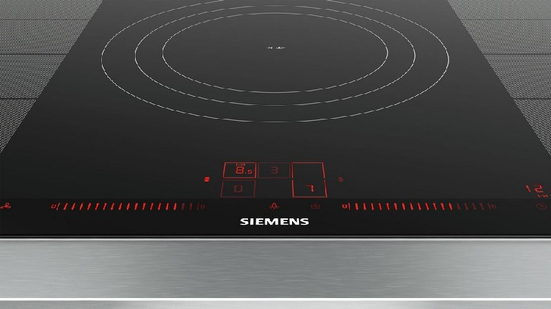 Primer plano de una placa de inducción Siemens. Superficie negra con números y controles iluminados en rojo. Marco de acero inoxidable.