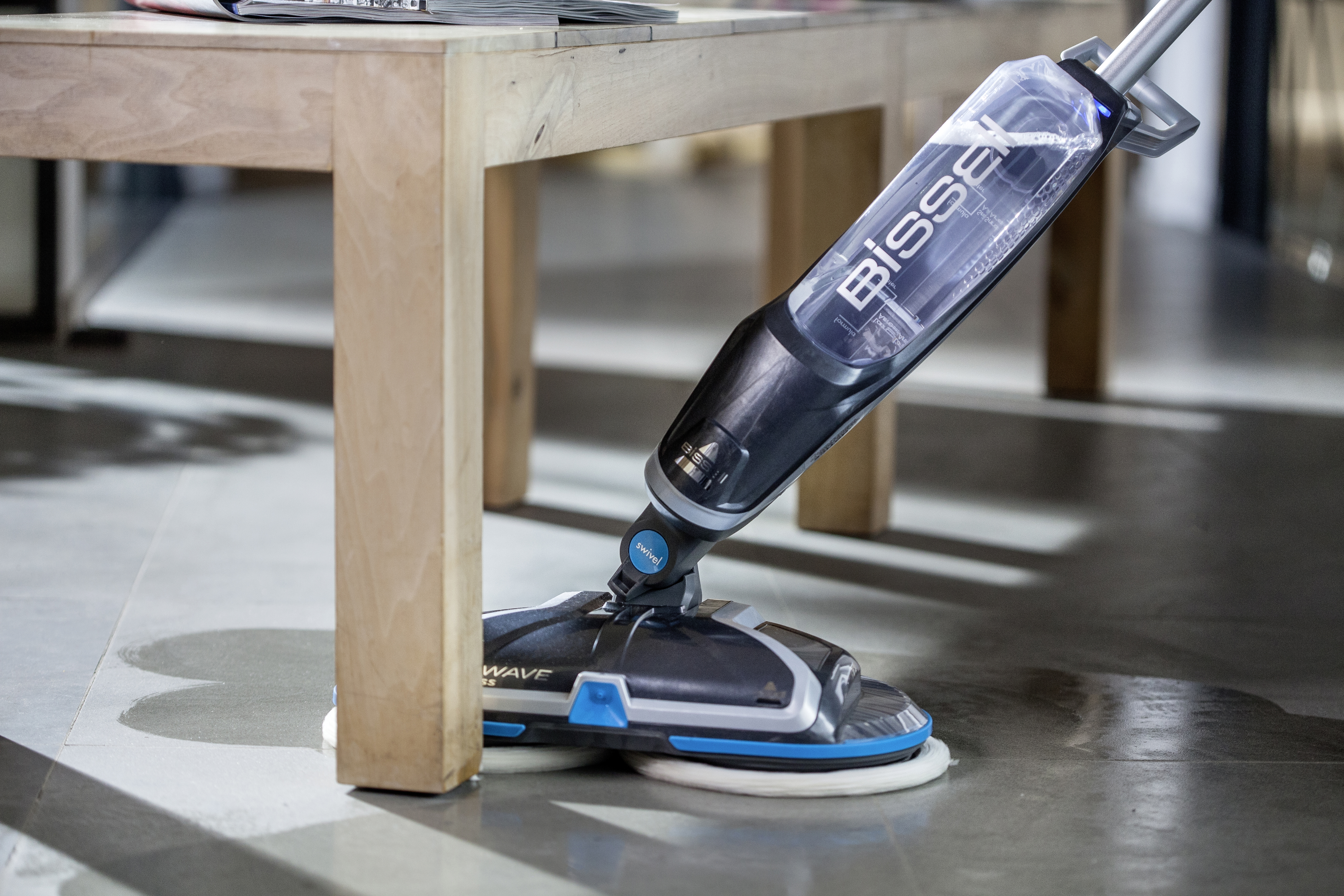 Bissell-Bodenreiniger in der Nähe eines Tischbeins. Schwarzes und blaues Gerät auf grauem Boden.