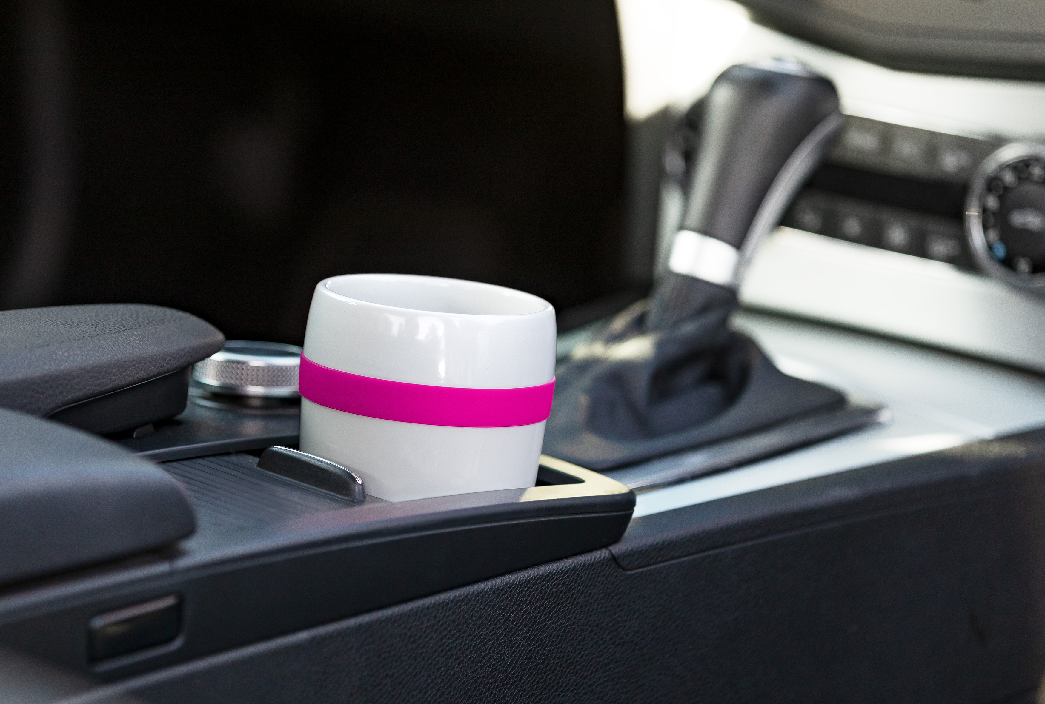 Ein weißer Becher mit rosa Band sitzt in einem Getränkehalter im Auto. Der Innenraum ist schwarz.