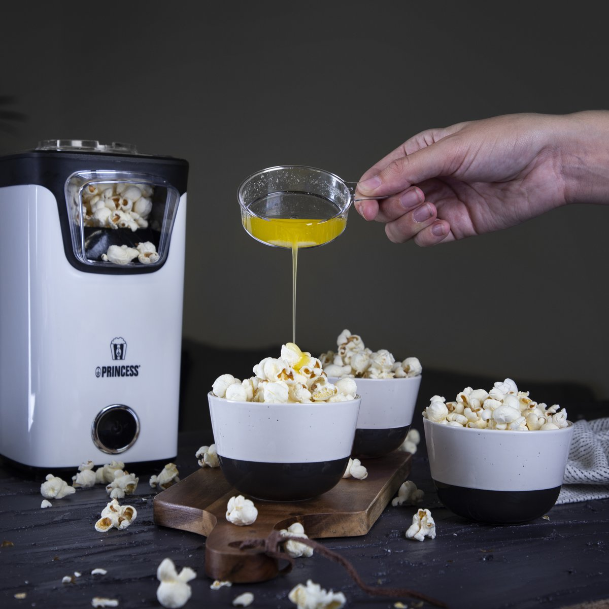 A person pouring yellow liquid onto popcorn in white and black bowls on wooden board.