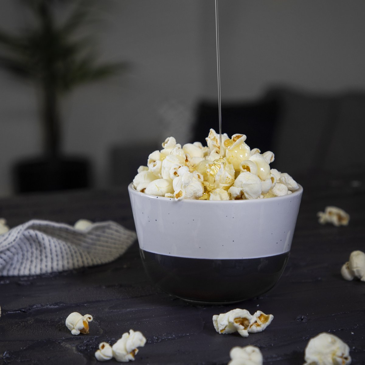 Bowl of popcorn with honey drizzled on top, on a dark table. Some popcorn scattered around.