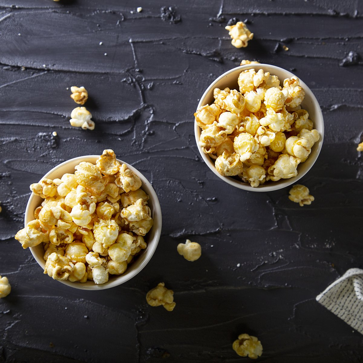 Two bowls of popcorn on a textured black surface. Some popcorn scattered around. A small white cloth in the corner.