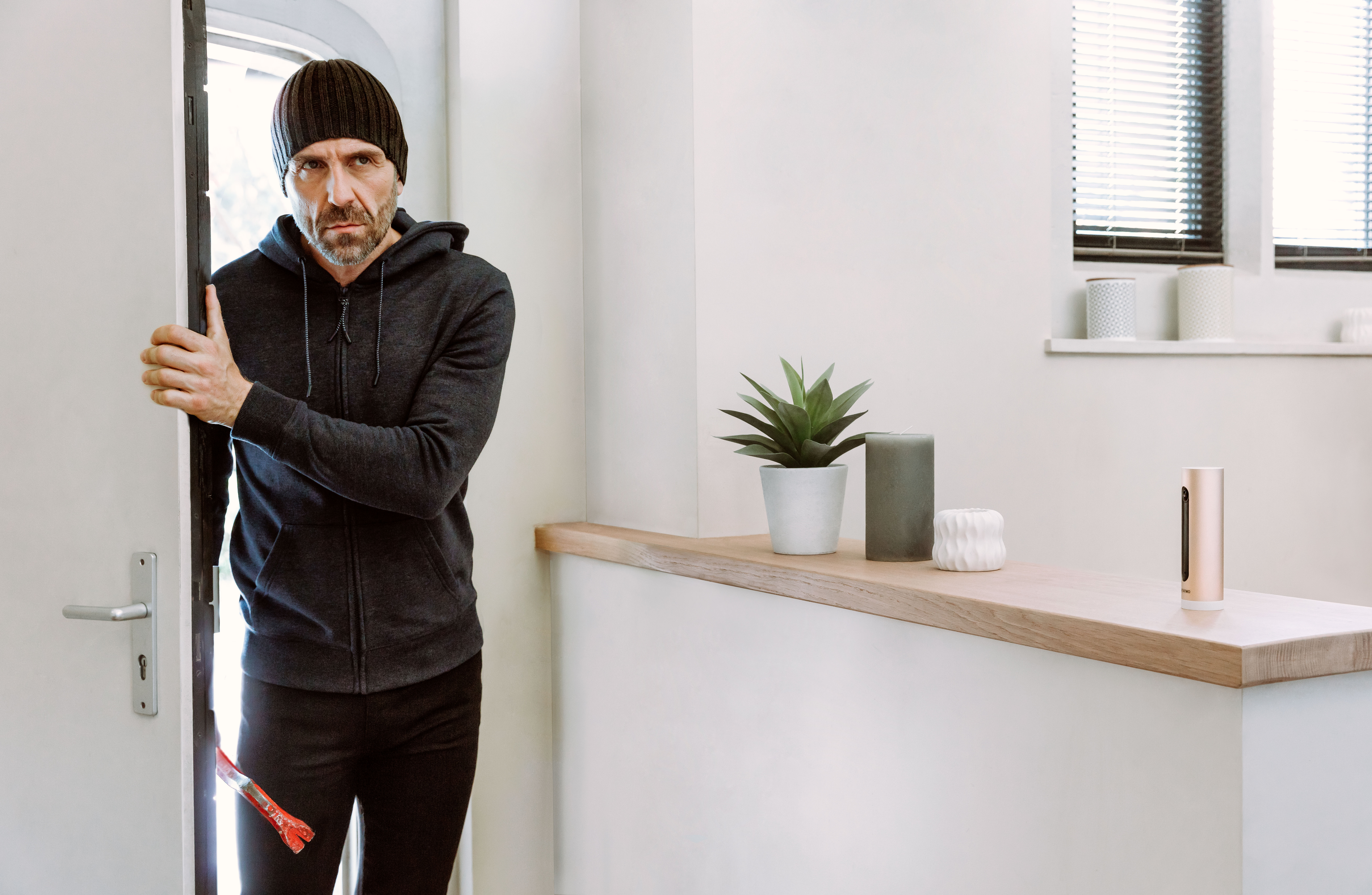 Homme en bonnet et vêtements sombres sortant d'une porte, tenant un pied-de-biche.