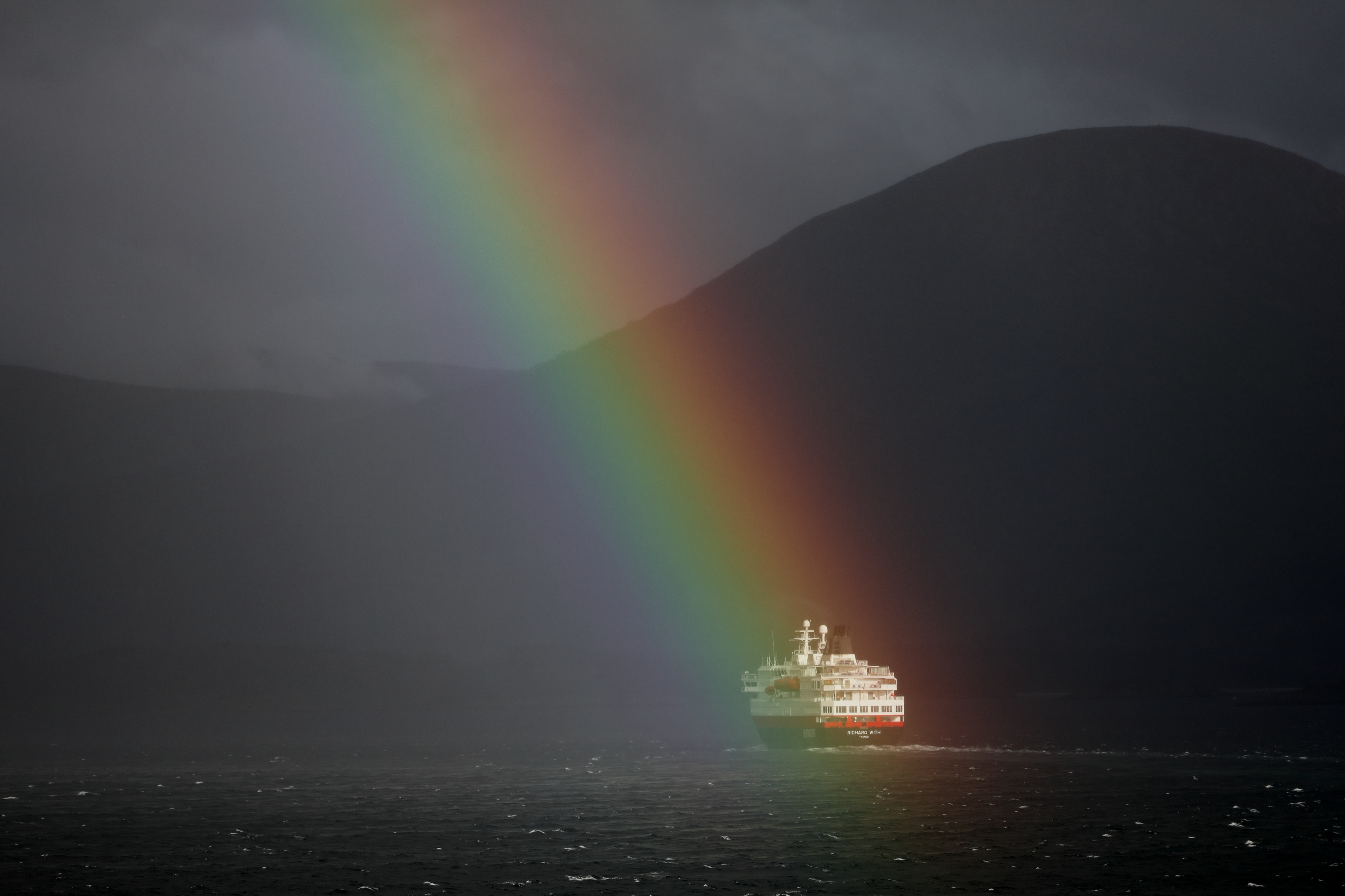 Regenbogen über Schiff auf Wasser, dunkler Berg im Hintergrund, stürmischer Himmel.
