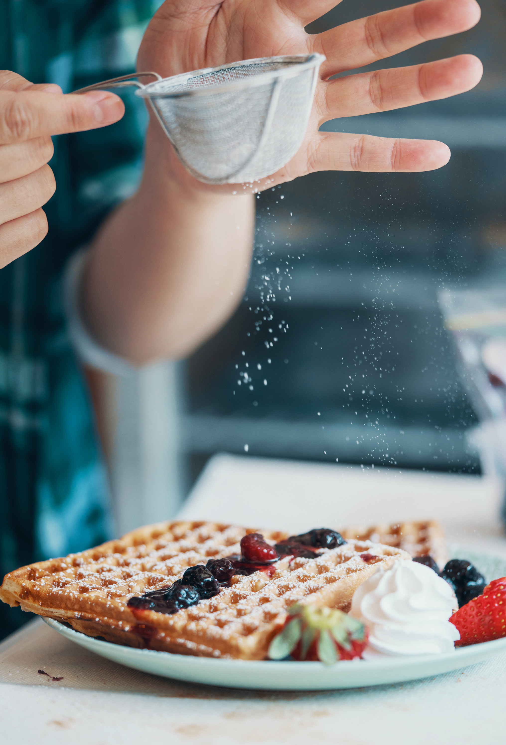 Puderzucker wird über Waffeln mit Beeren und Schlagsahne auf einem Teller gestreut.