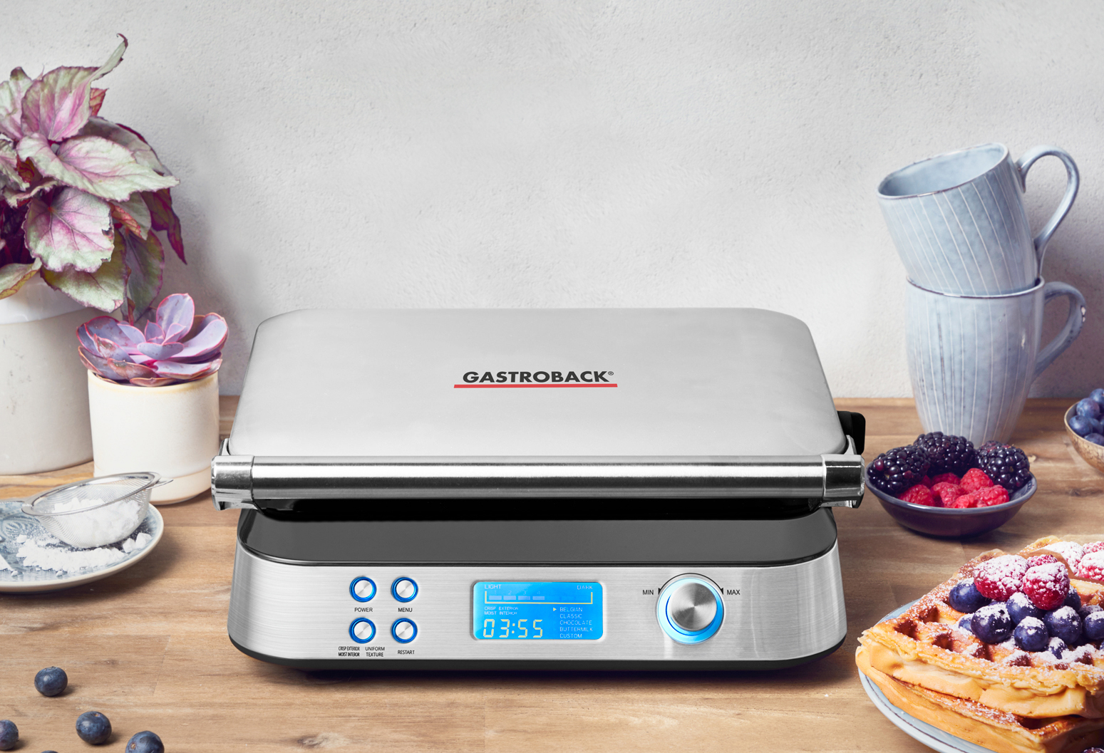 Silber-schwarzer Waffeleisen mit Digitalanzeige. Beeren und Waffeln an der Seite. Holztisch.