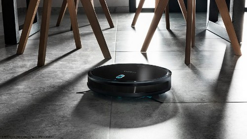 Aspiradora robot negra en un suelo de baldosas grises. Patas de mesa de madera al fondo.