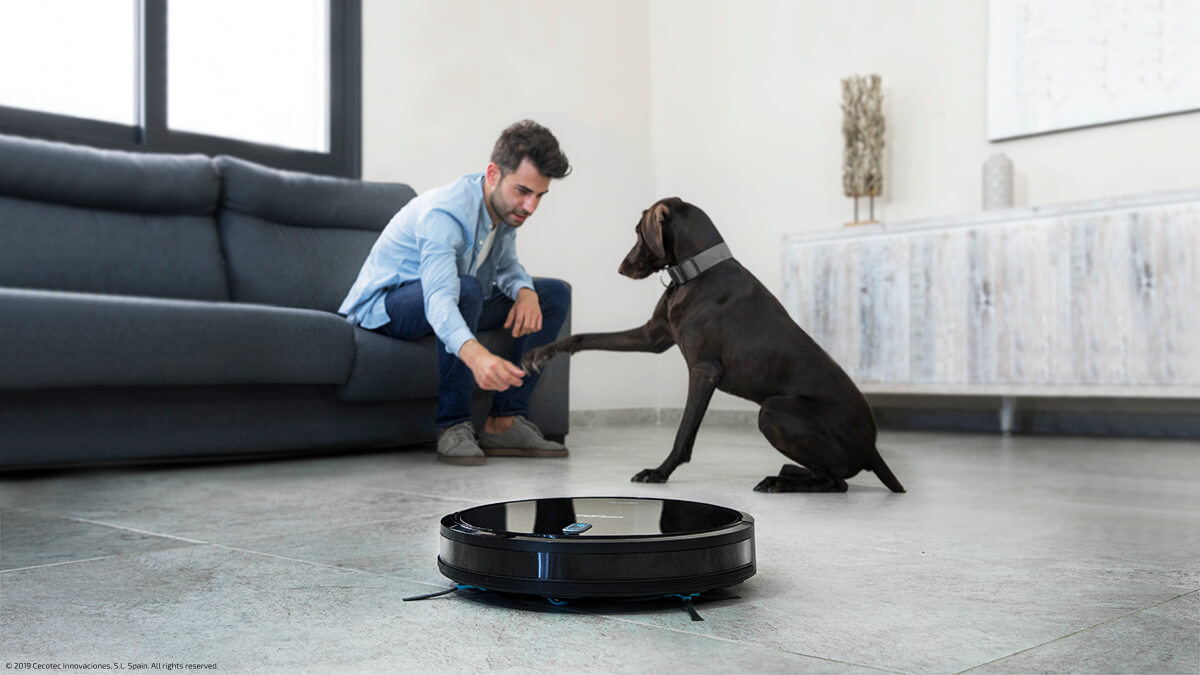 Un hombre y un perro interactuando con un robot aspirador en el suelo.
