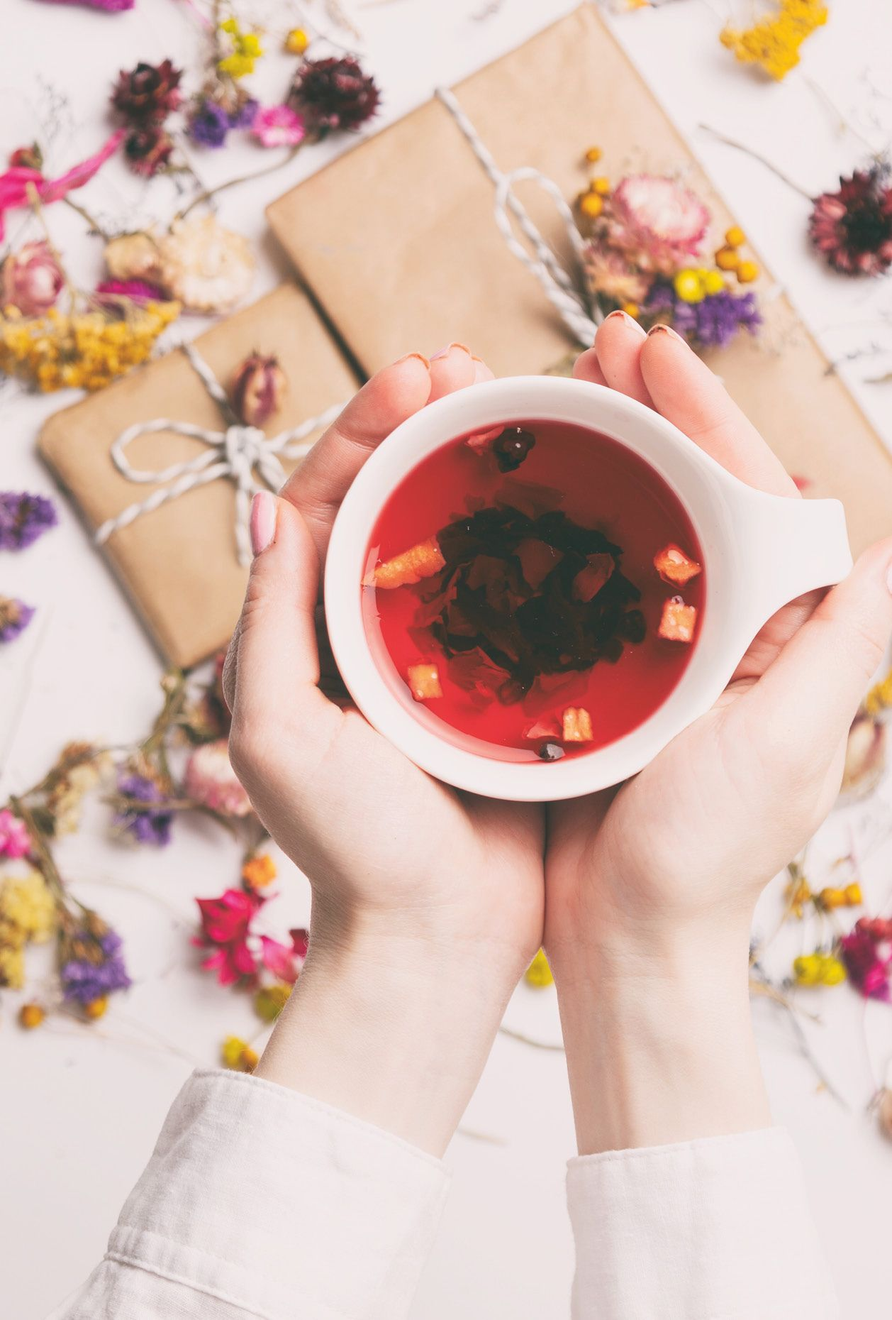 Hände halten eine weiße Tasse mit rotem Tee. Geschenke und Blumen sind im Hintergrund.