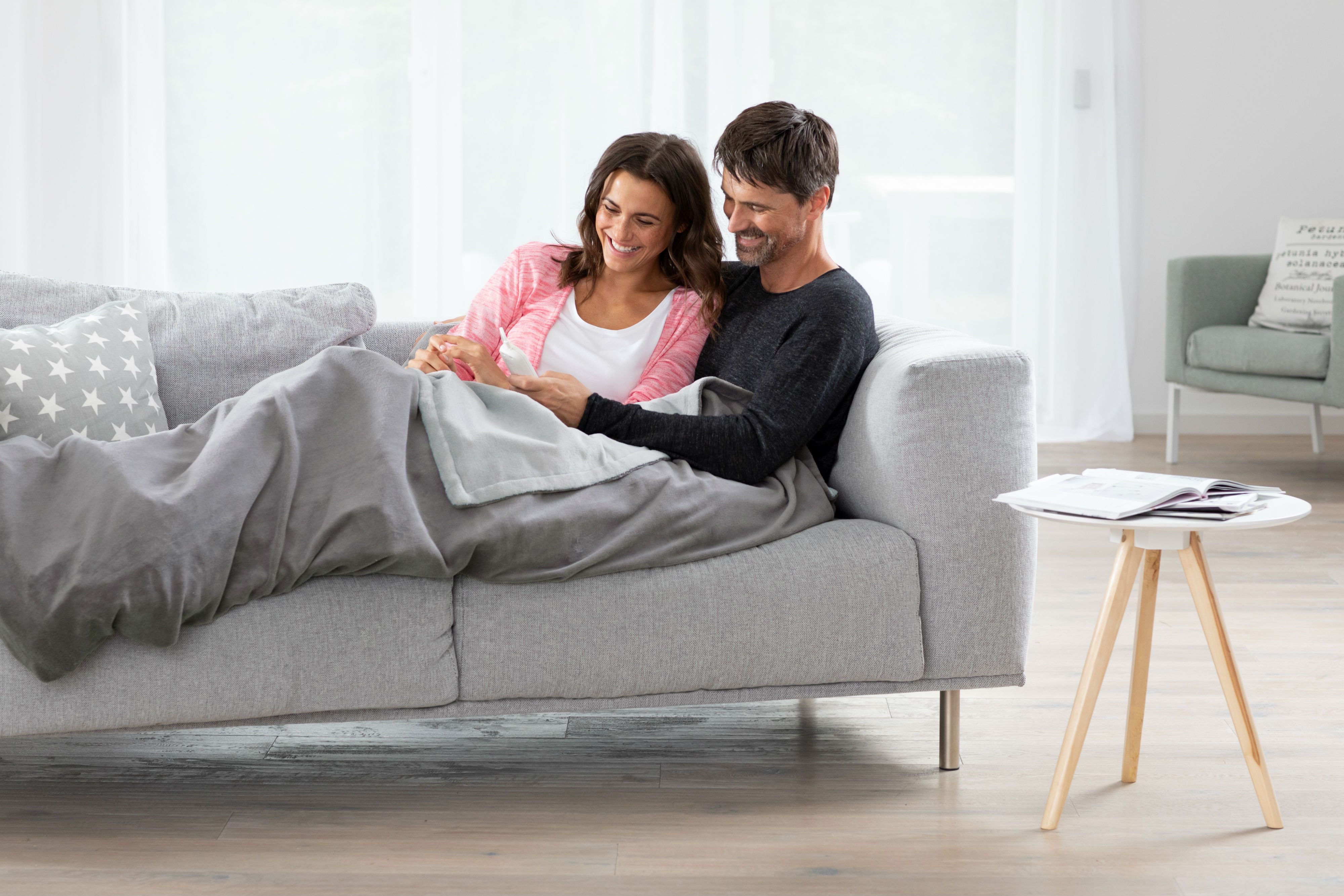 Paar auf einer grauen Couch, schauen auf ein Handy. Eine Decke bedeckt sie; ein Beistelltisch ist daneben.