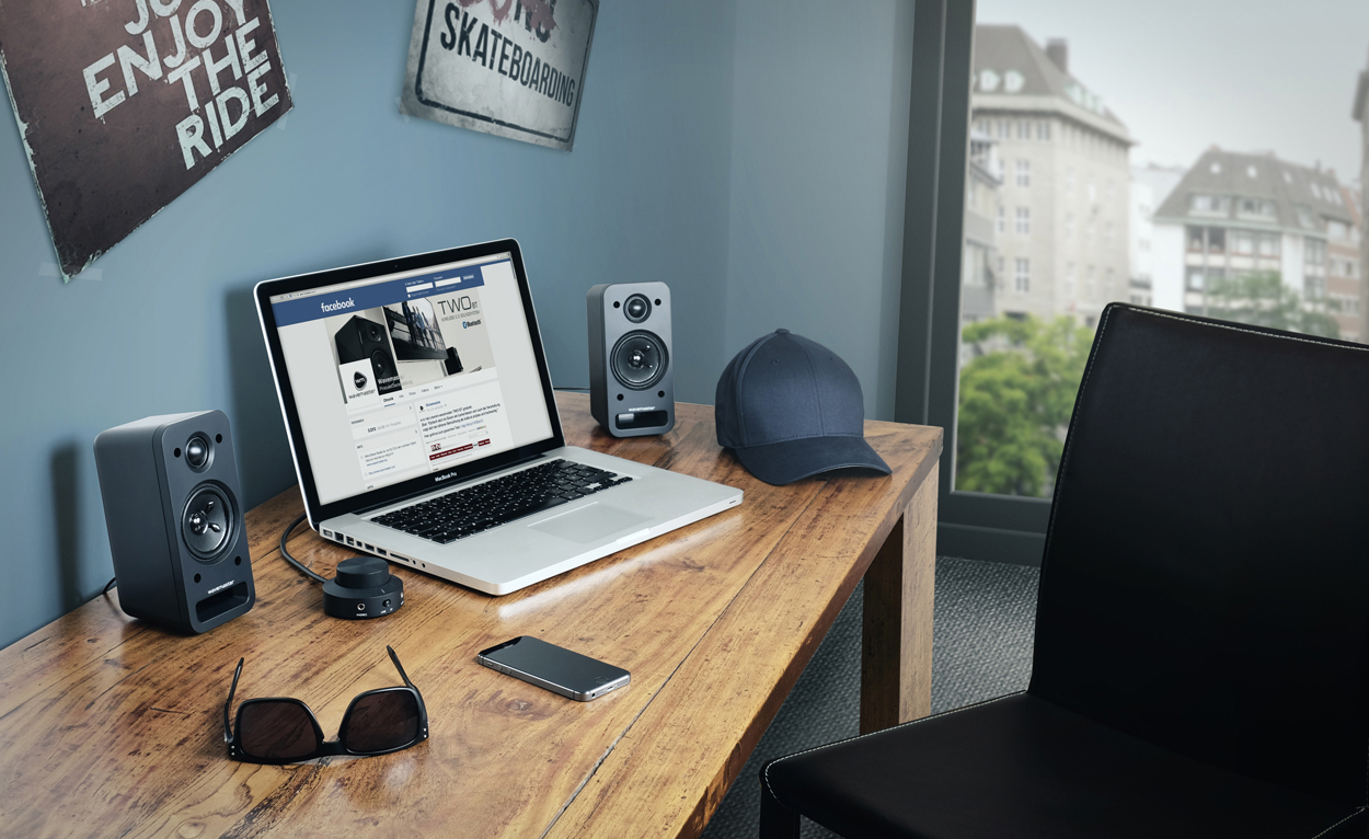 Laptop auf einem Schreibtisch mit Lautsprechern, Sonnenbrille und Mütze. Hintergrund ist ein Raum.