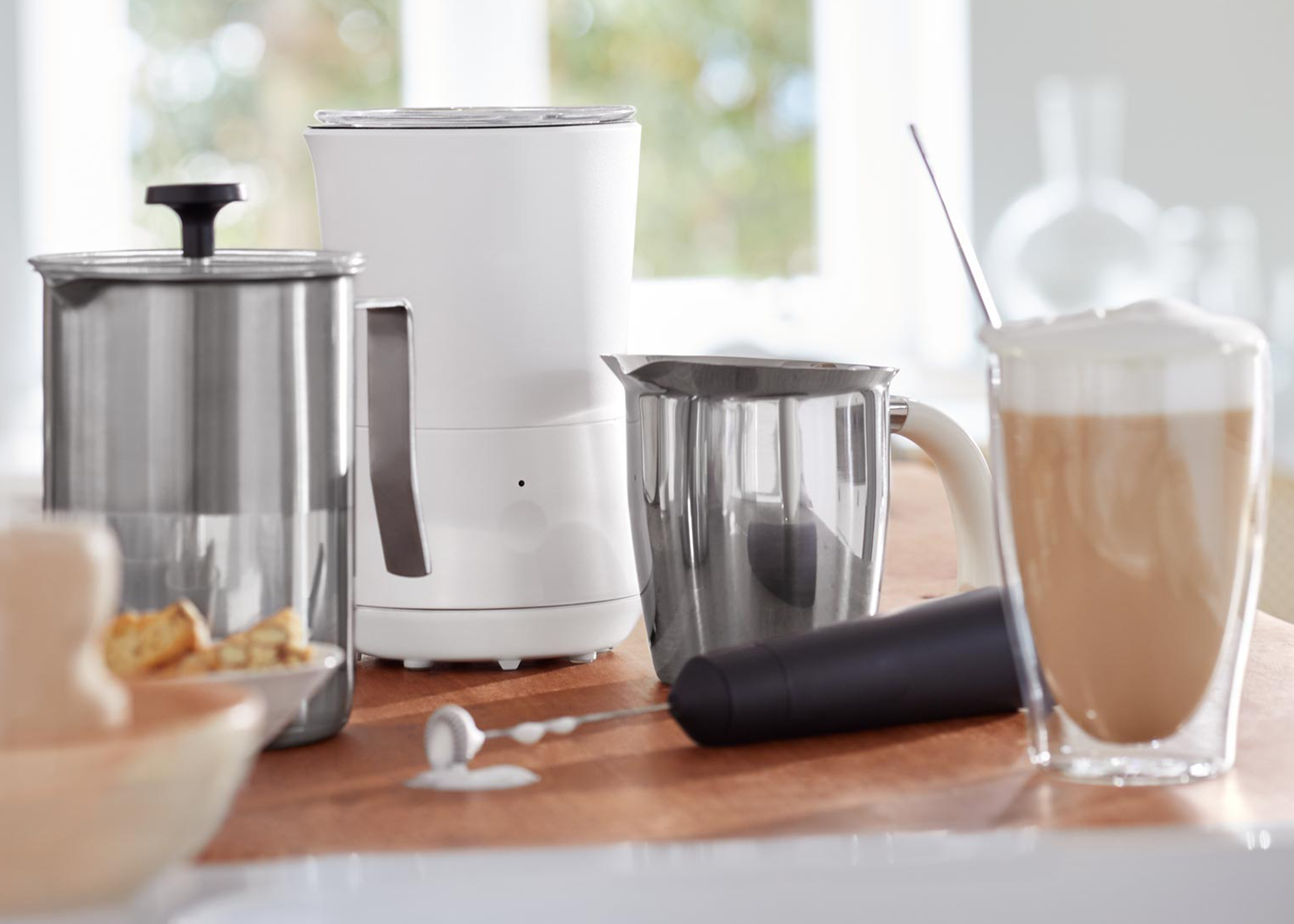 Küchenszene mit Kaffeemaschine, Milchaufschäumer, Kaffee und einem Löffel. Szene auf Holzoberfläche.