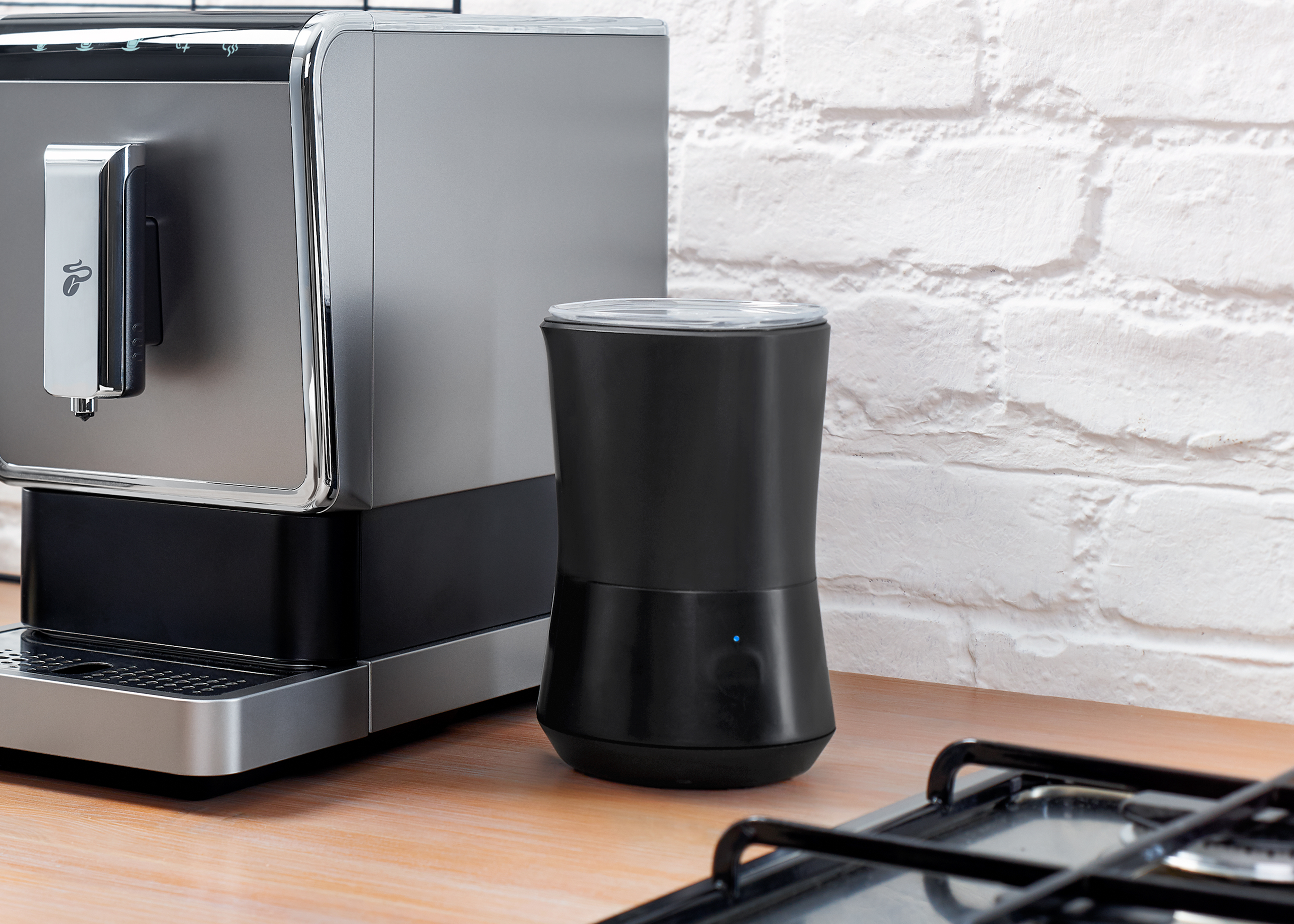 Kaffeemaschine mit Milchaufschäumer. Das Setup befindet sich auf einer Holzoberfläche in der Nähe einer Ziegelmauer.