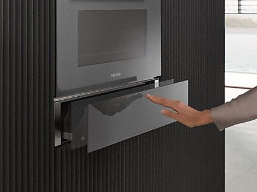 Eine Hand öffnet eine Schublade unter einem Miele-Ofen mit dunkler Wand.