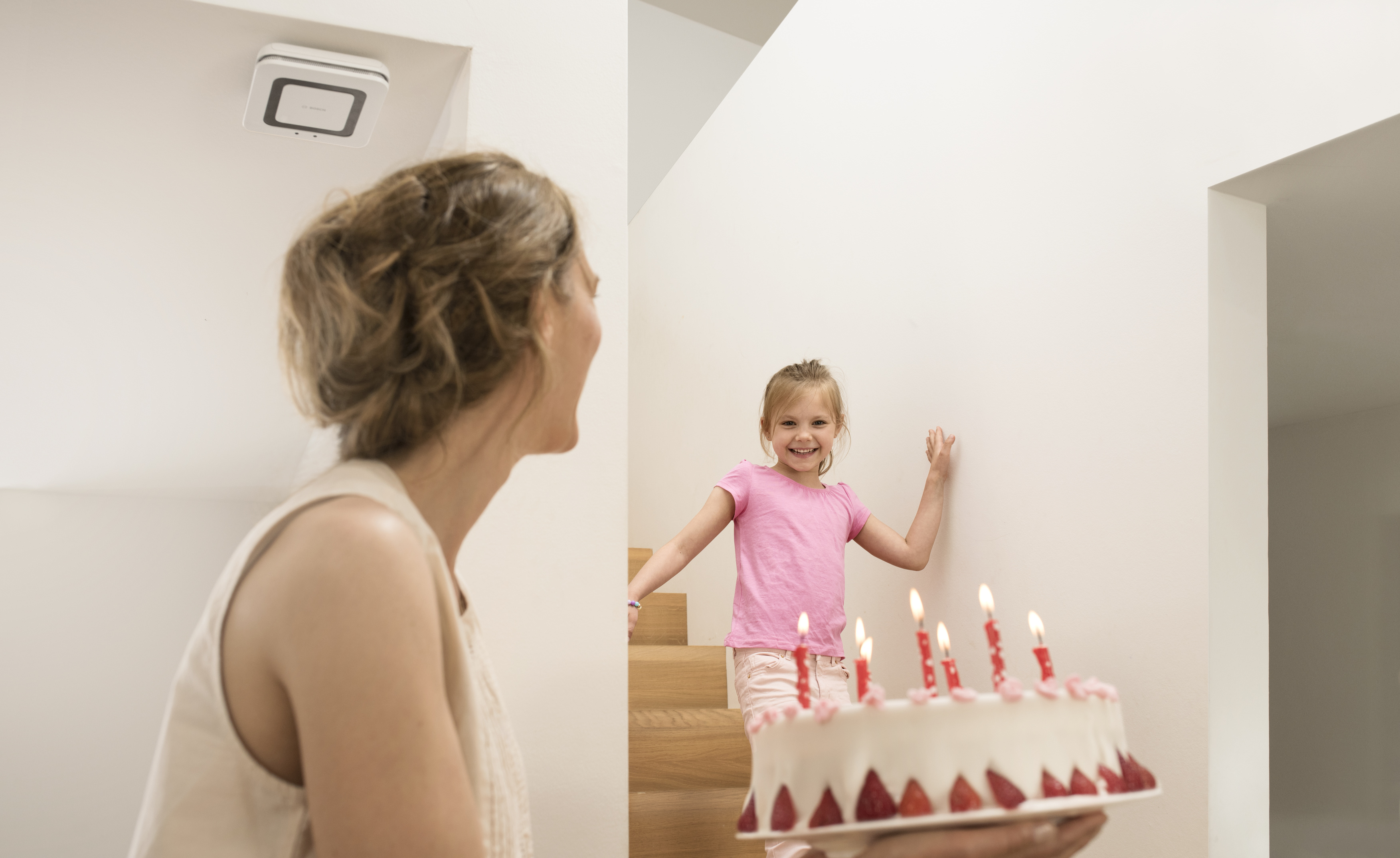 Eine Frau schaut ein Mädchen an, das einen Geburtstagskuchen mit Kerzen hält, Rauchmelder im Hintergrund.