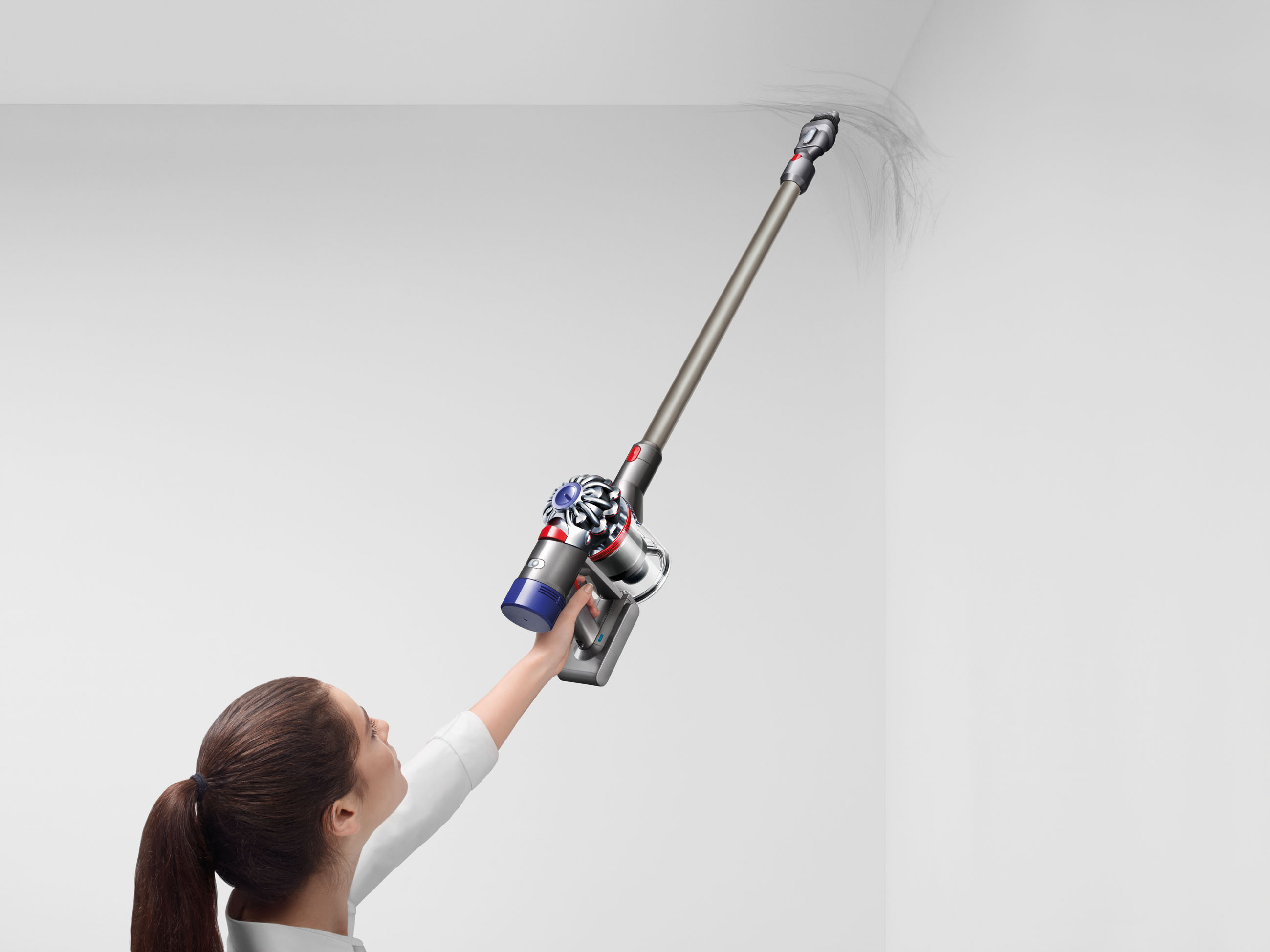 Eine Frau benutzt einen silbernen und roten Staubsauger, um eine weiße Wand zu reinigen. Staub ist sichtbar.