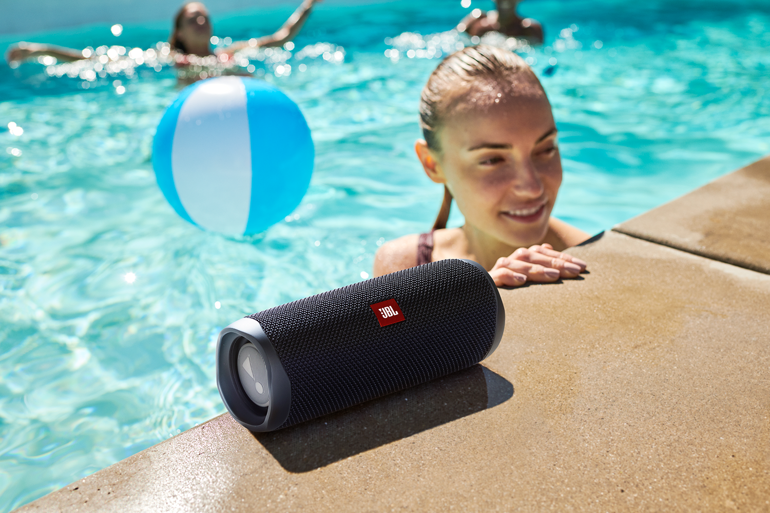 Een zwarte JBL-luidspreker staat bij een zwembad. Een vrouw glimlacht en een blauw-witte strandbal drijft in het water.